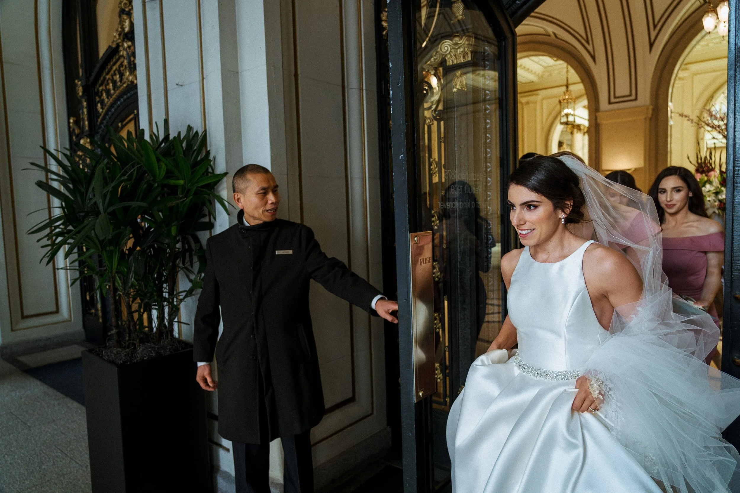 A bride in a white wedding dress exiting a building with a beaming smile, holding her gown, as a doorman opens the door for her. Two women in pink dresses follow behind.