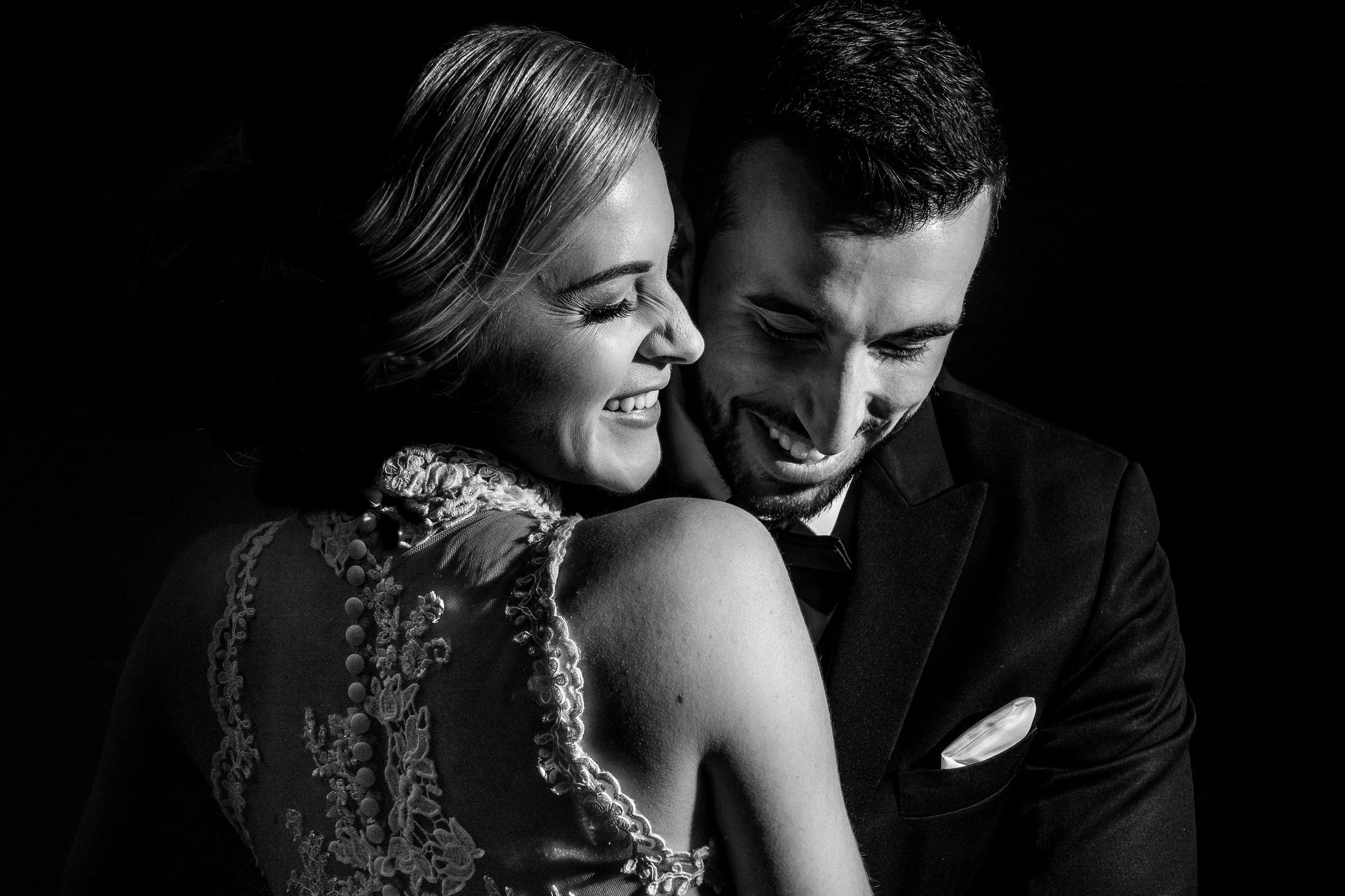 A black and white photo of a smiling bride and groom close together, with the bride in a lace dress and the groom in a tuxedo, sharing a joyful moment.