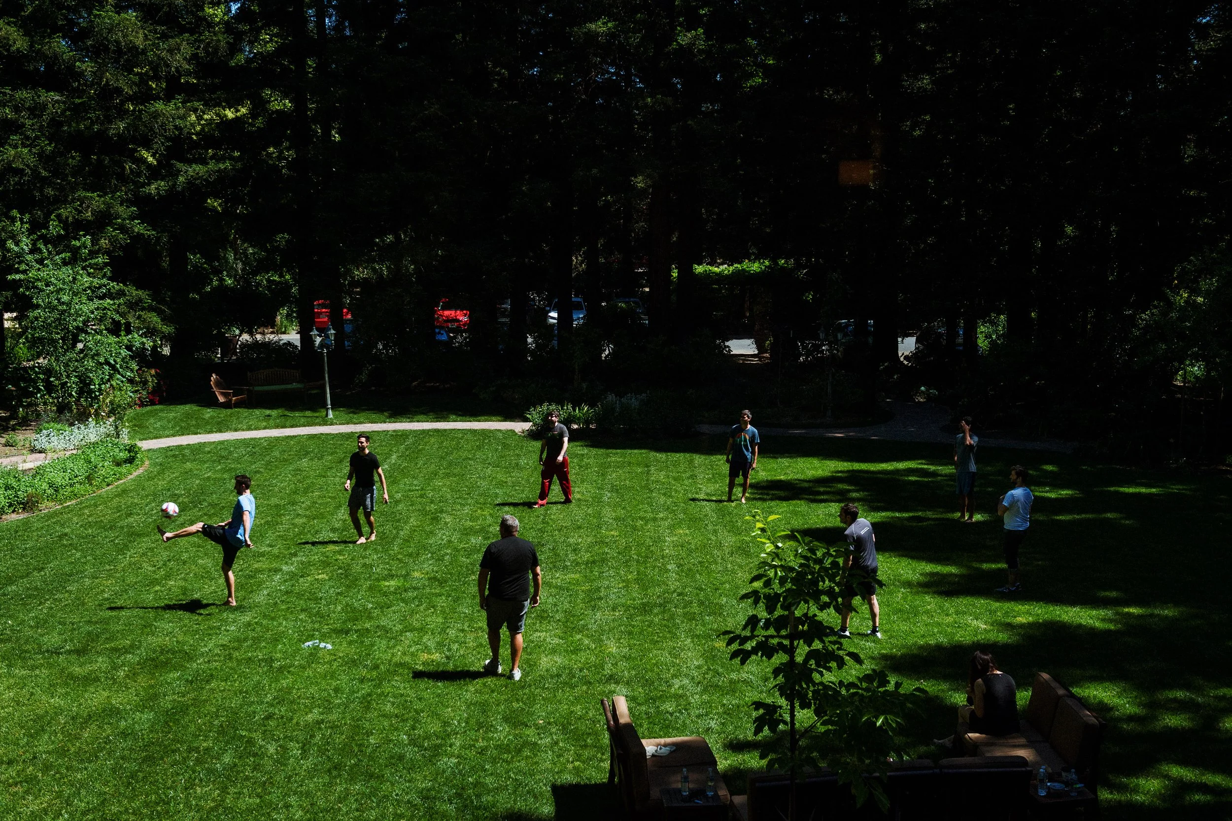 People playing soccer in a park on a grassy field, surrounded by trees and shaded areas.