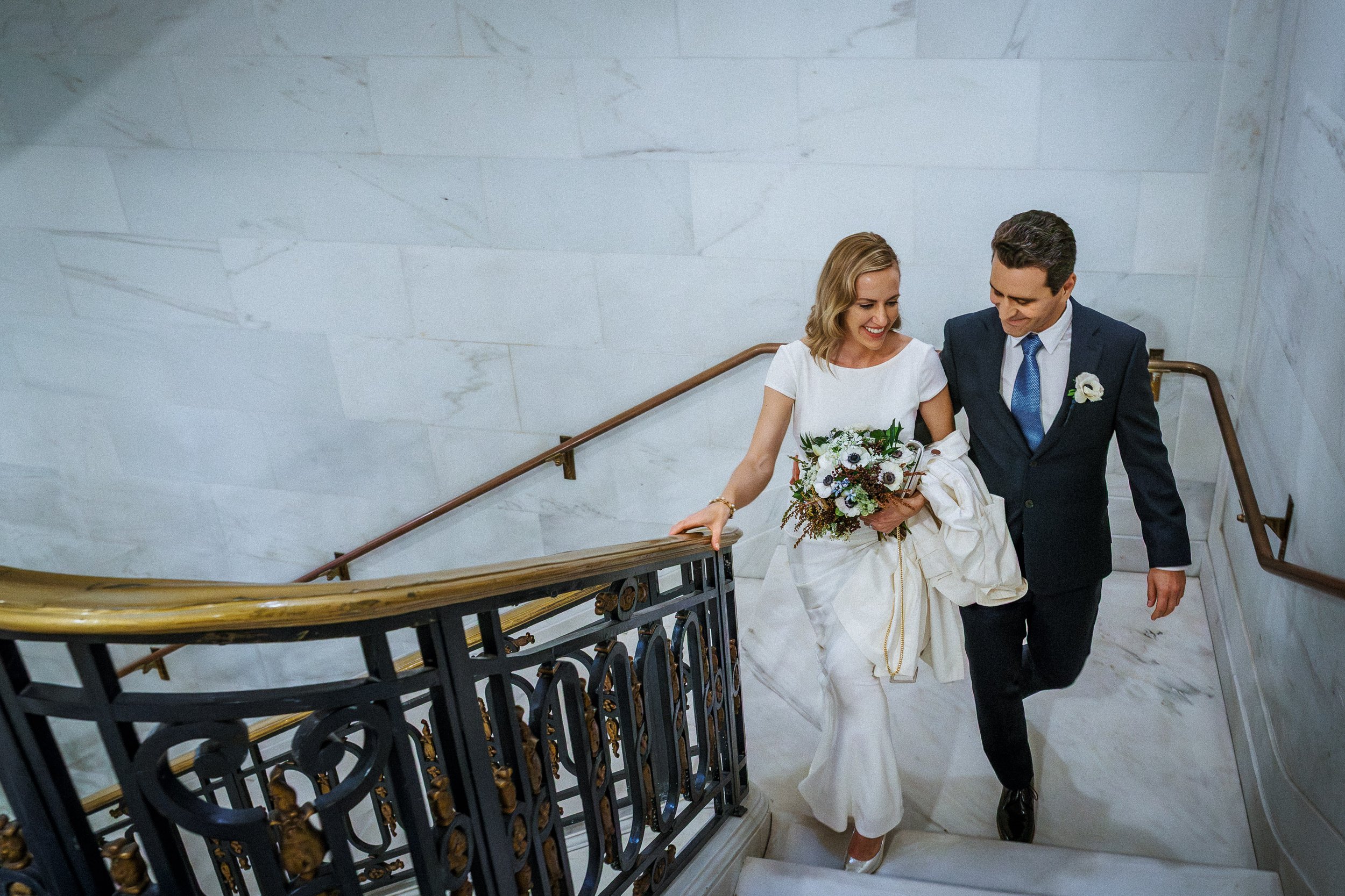 A bride in a white dress holding a bouquet, descending a marble staircase with a groom in a dark suit, both smiling.