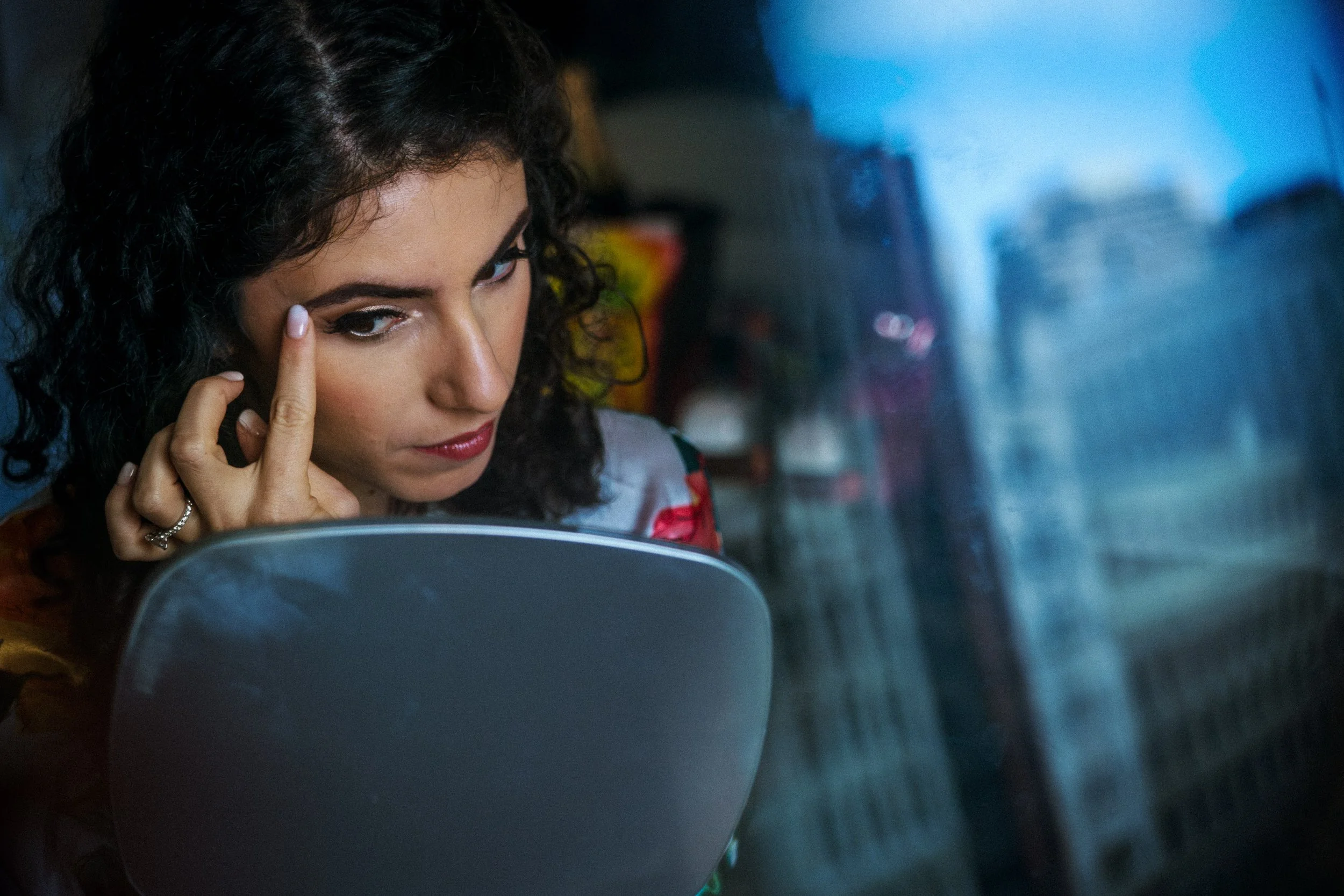 A woman with curly dark hair applying makeup or eyeliner while looking into a mirror, in a dimly lit room at the Palace Hotel in San Francisco, with blurred background objects.
