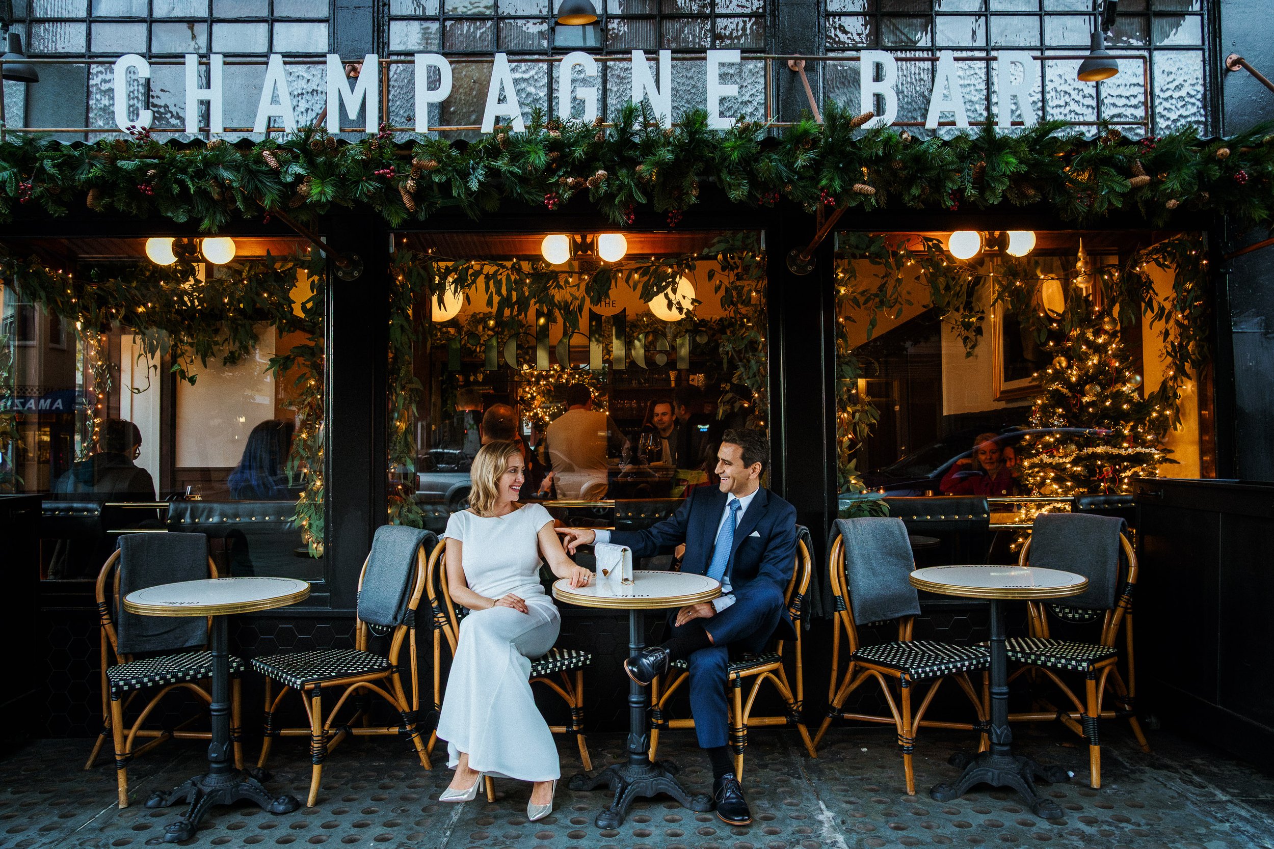 A couple sitting outside a champagne bar, smiling and holding hands, with festive holiday decorations and a Christmas tree inside.
