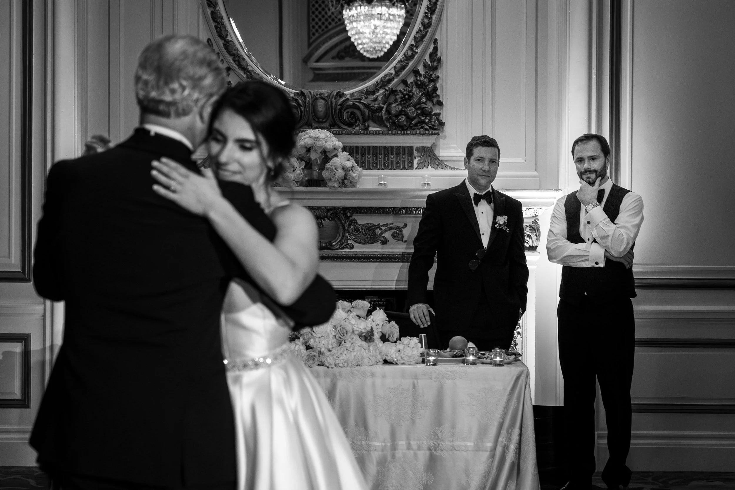 A black and white photo of a wedding reception. The newlyweds are dancing, with the bride smiling as she embraces the groom who is facing away from the camera. Two men in formal tuxedos are standing near a decorated table, watching the couple, with o