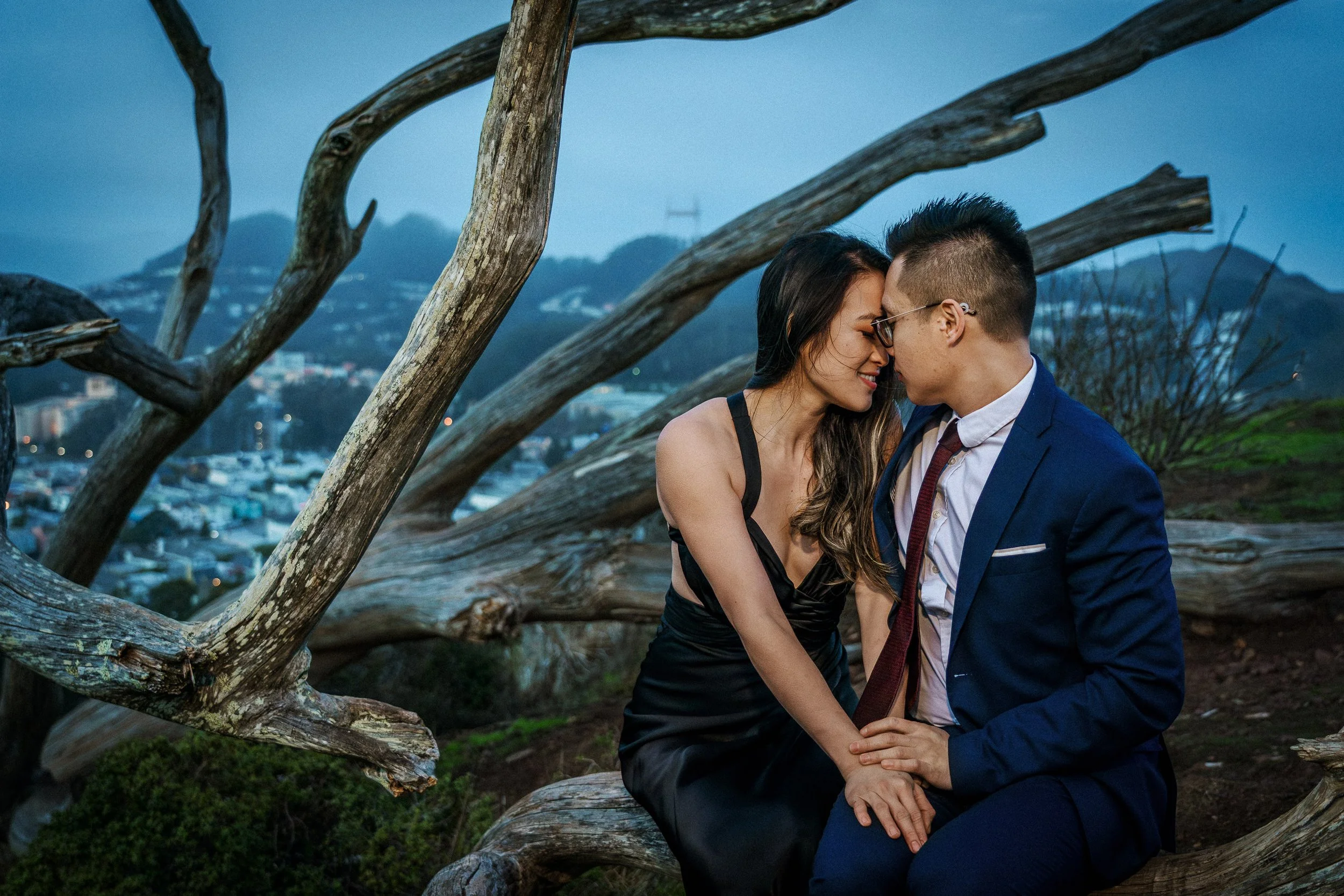 A couple sitting close together on a fallen tree trunk outdoors during evening, nose to nose, with the San Francisco cityscape and mountains in the background.