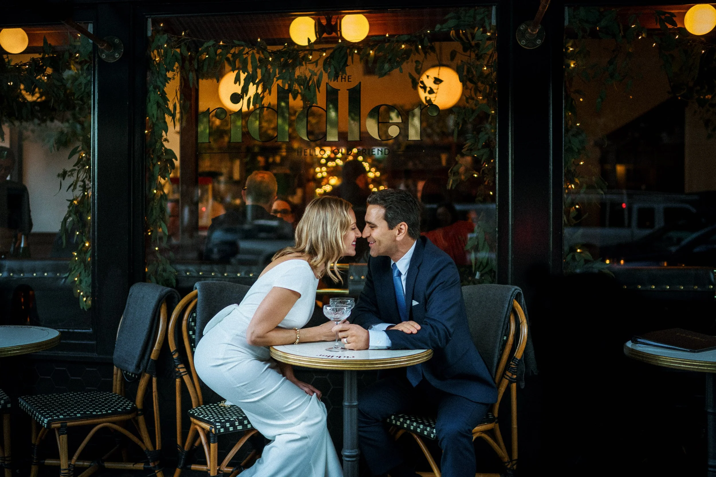 Couple having an intimate romantic moment inside a dimly lit restaurant. The bride in a white dress and the groom in a dark suit lean close, smiling and holding a champagne glass.