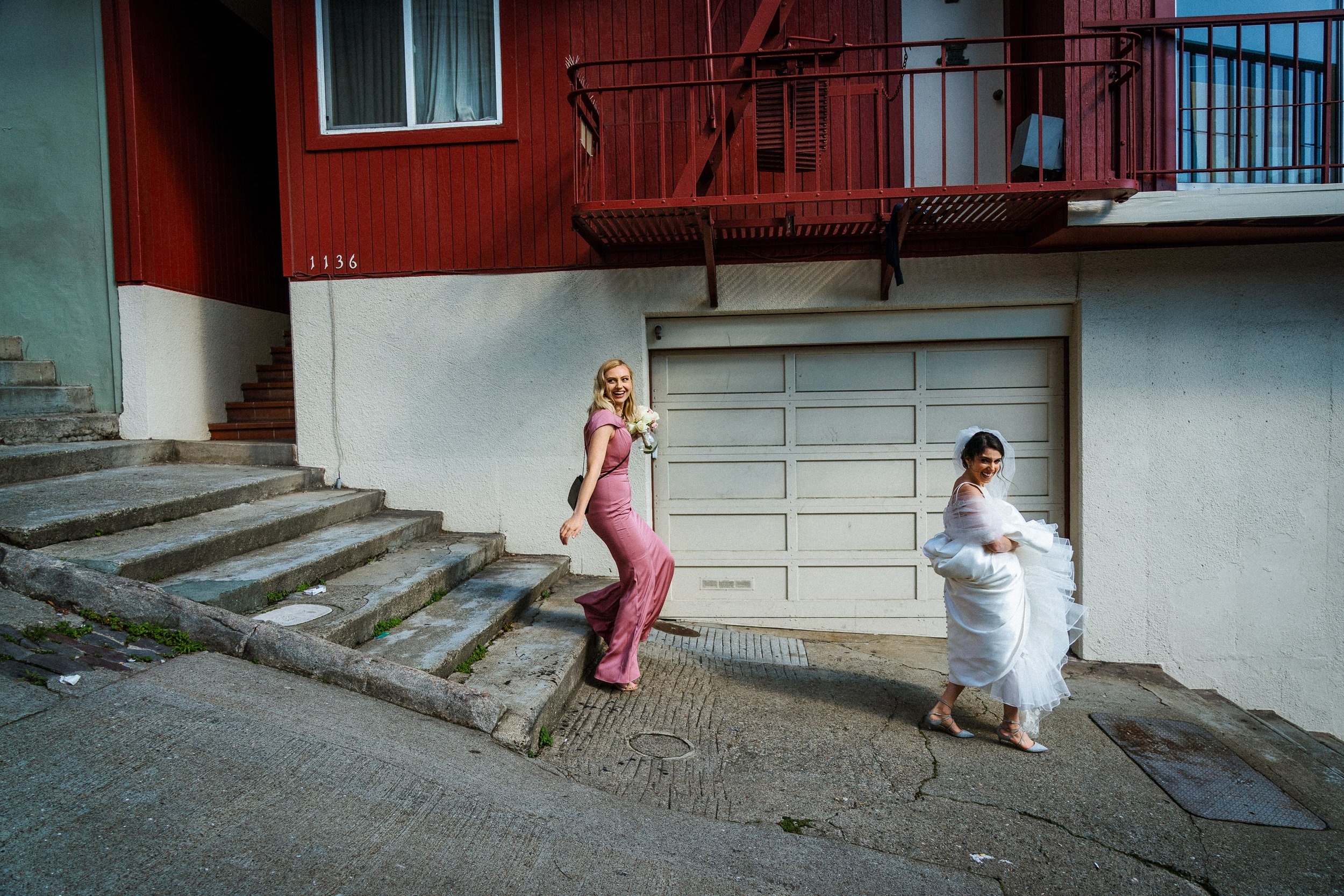 Two women, one in a pink bridesmaid dress and the other in a wedding dress, are walking on a street in front of a building with a garage door, stairs, and a red upper facade.