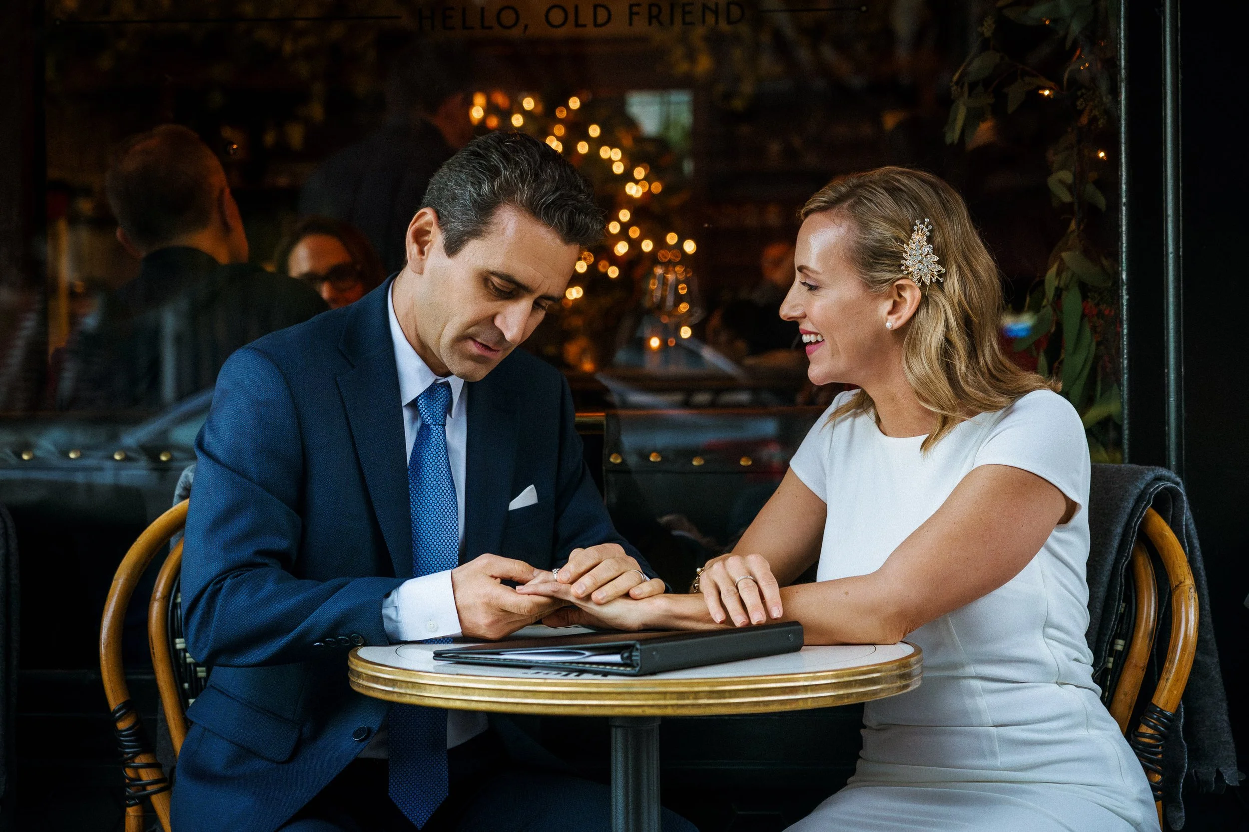 A man in a blue suit and a woman in a white wedding dress sitting at a table in a restaurant, with the woman smiling happily. The background is decorated with warm lighting and a Christmas tree.