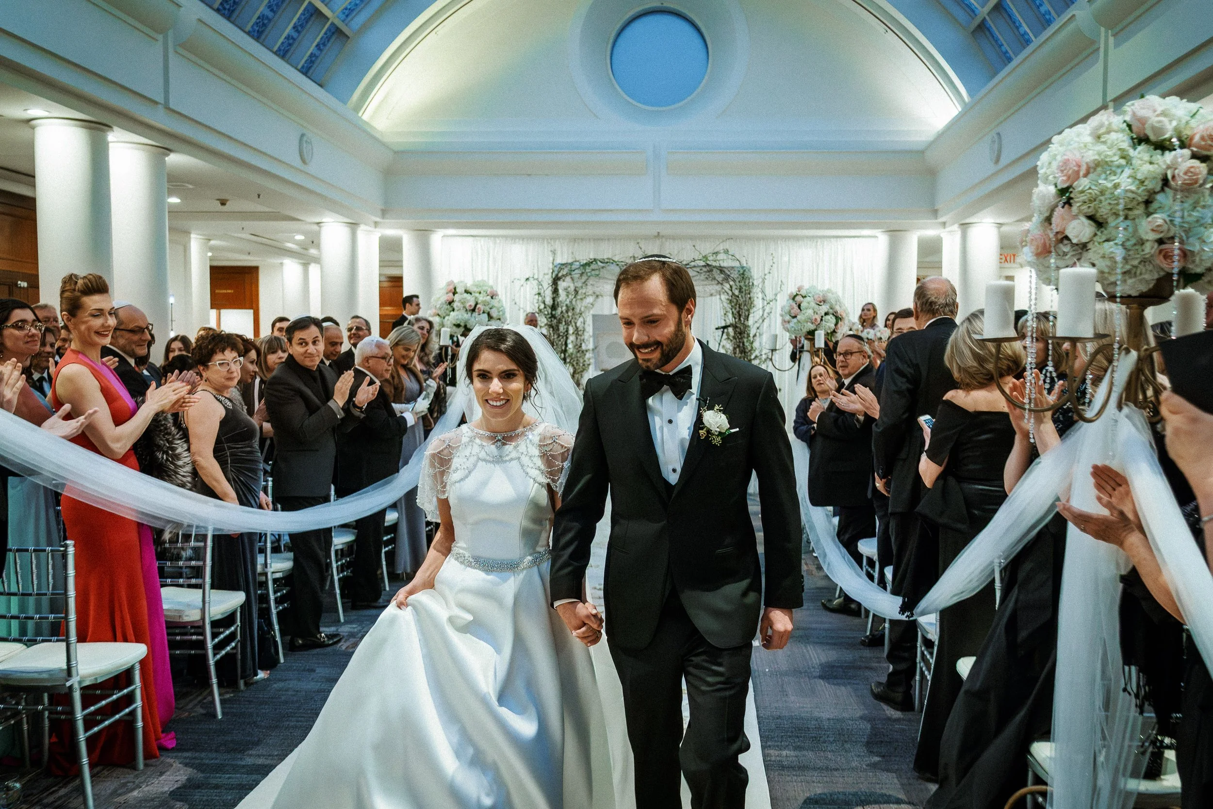 A bride and groom walking down the aisle at their wedding ceremony in a decorated indoor venue, surrounded by clapping guests.
