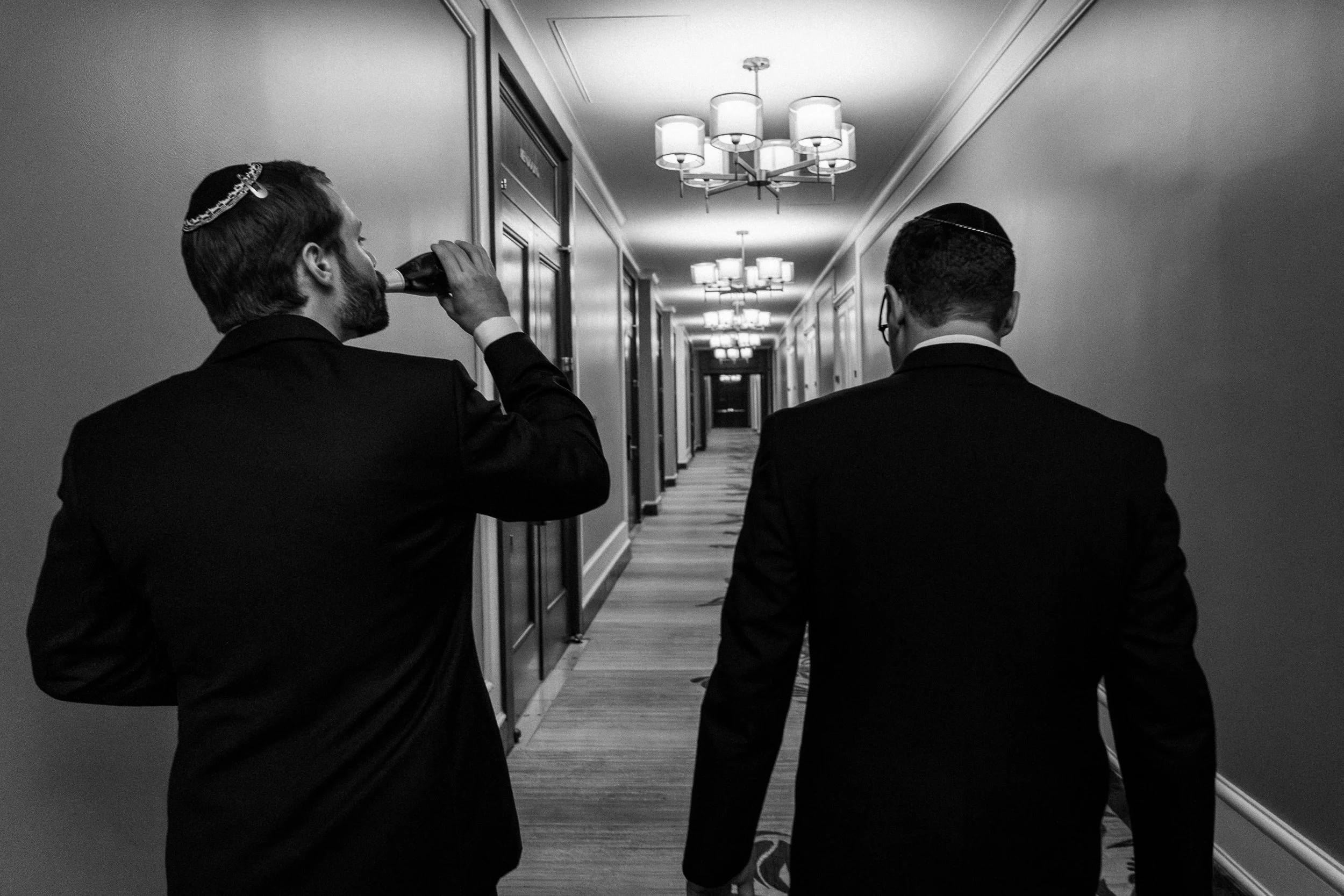 Two men in suits and kippahs walking down a hotel corridor, one drinking from a bottle.