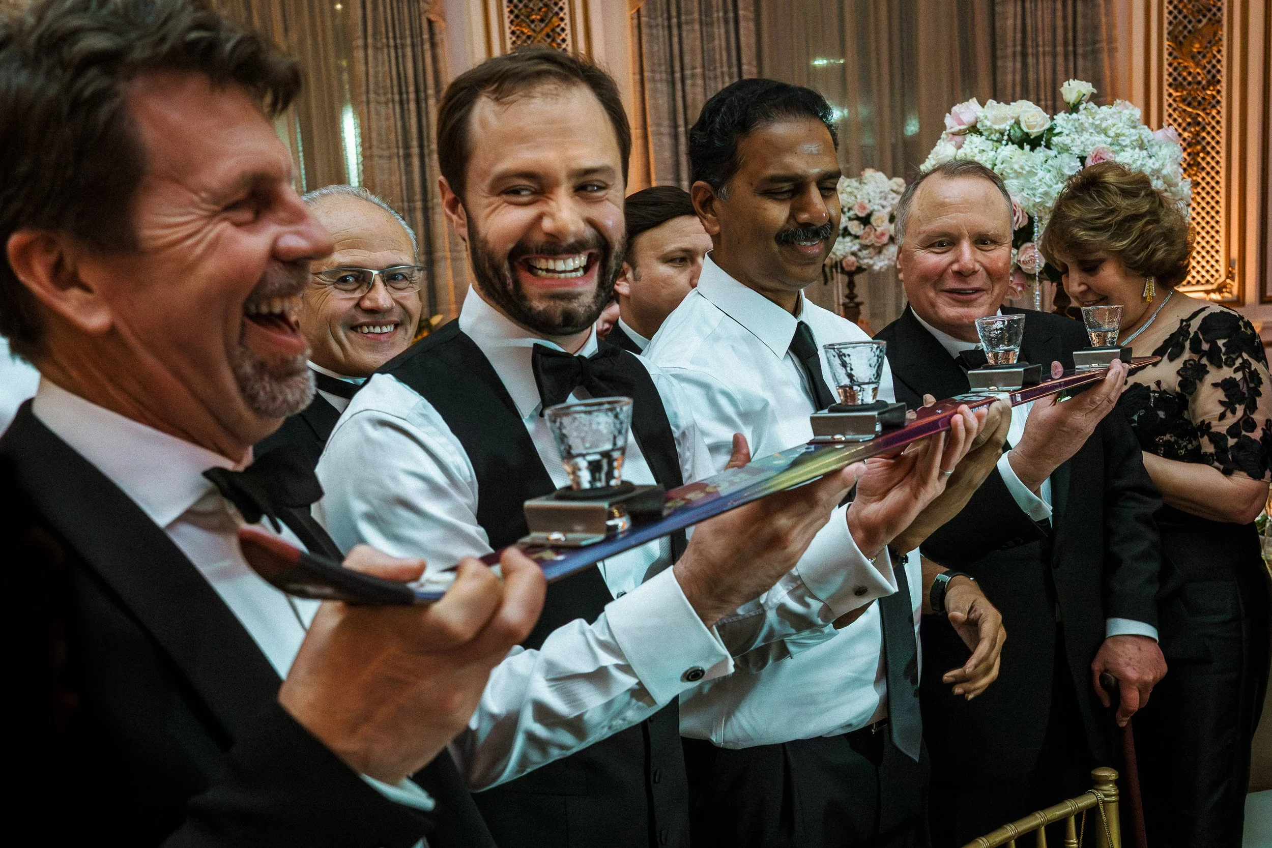 A group of people dressed in formal attire, participating in a wine tasting event at a celebration or wedding, with a floral centerpiece in the background.
