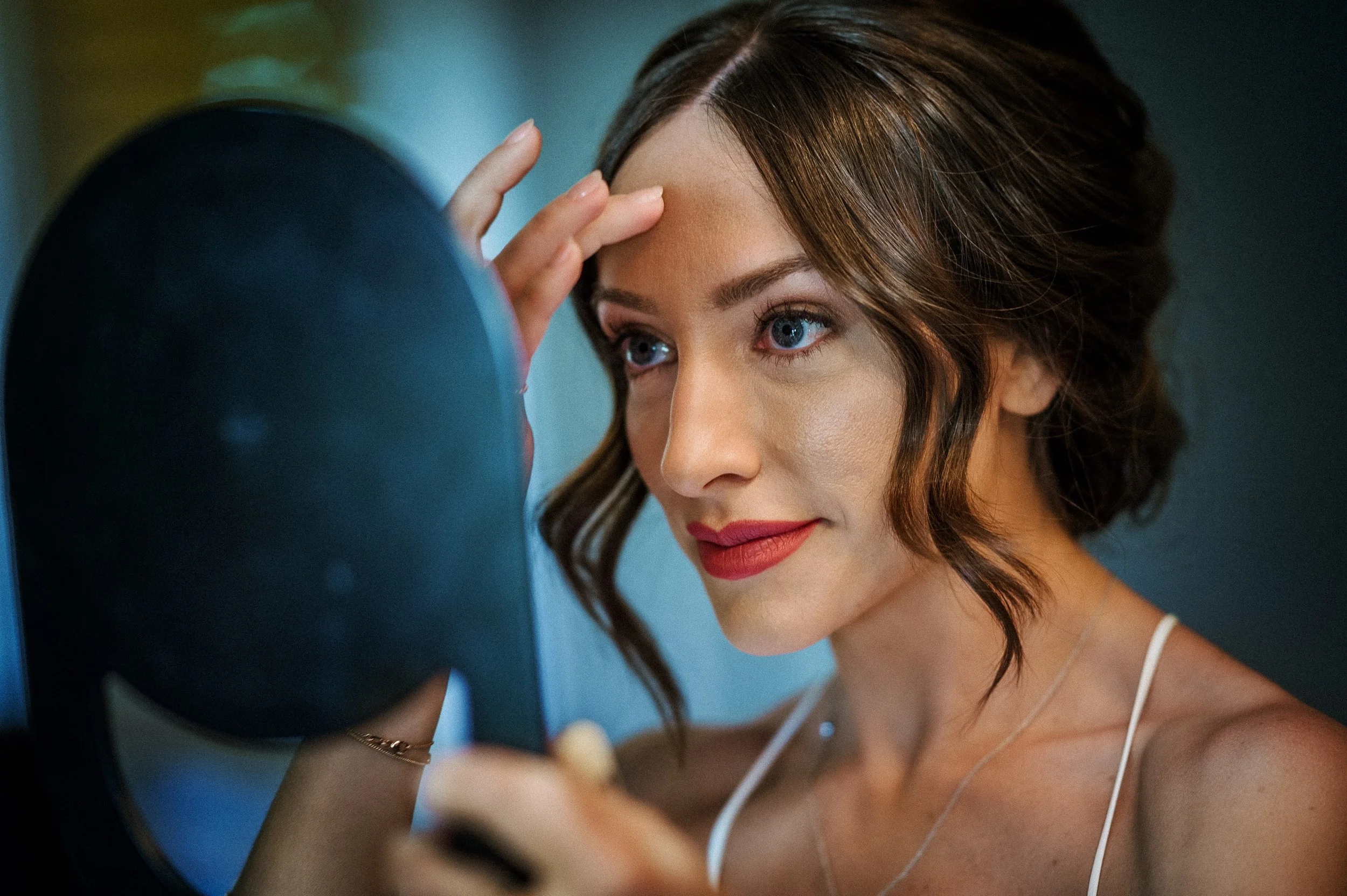 A bride with wavy brown hair and blue eyes looking in a mirror while applying makeup.