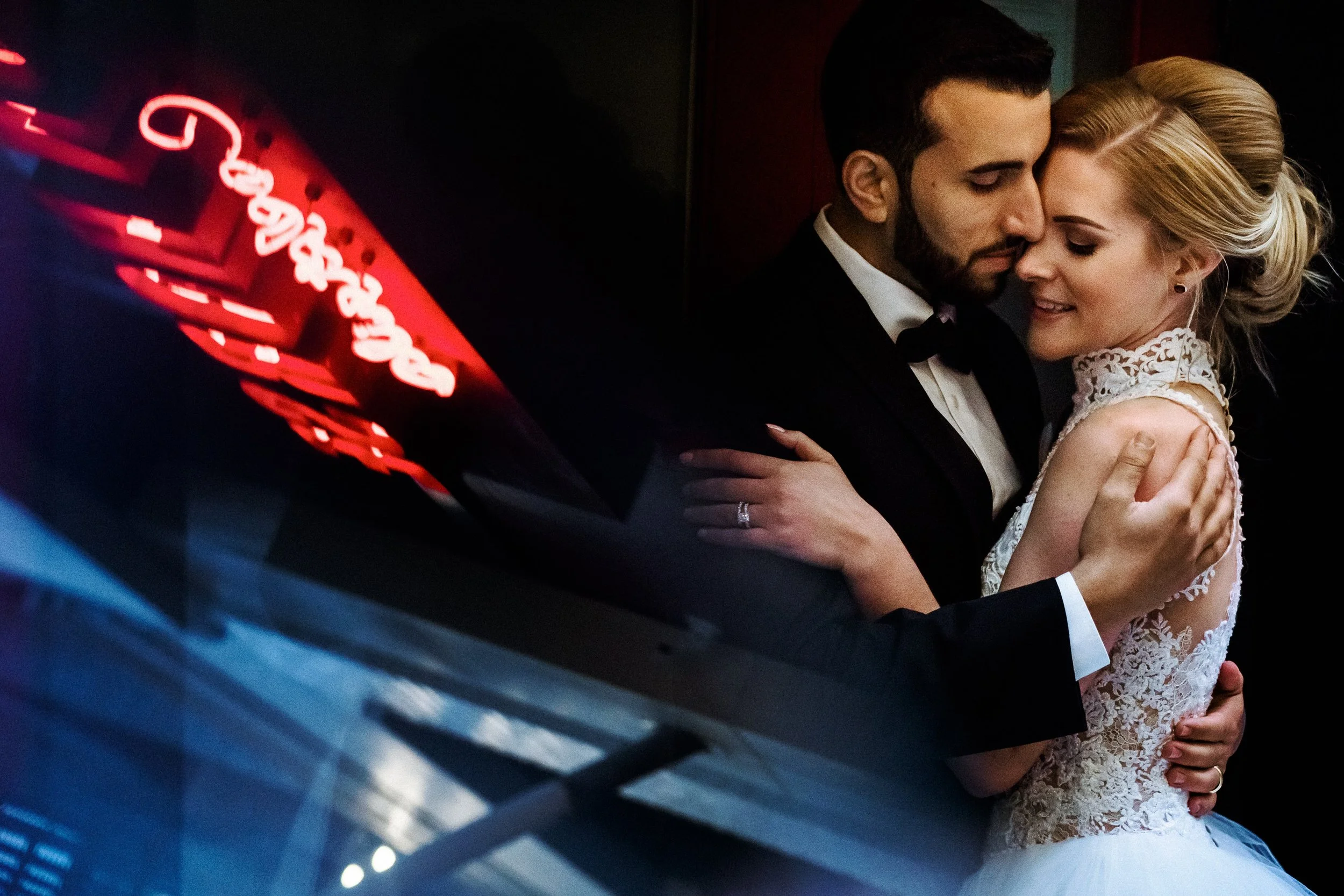 A newlywed couple embracing each other, the groom in a tuxedo and the bride in a lace gown, with a neon sign reflected in a nearby window.