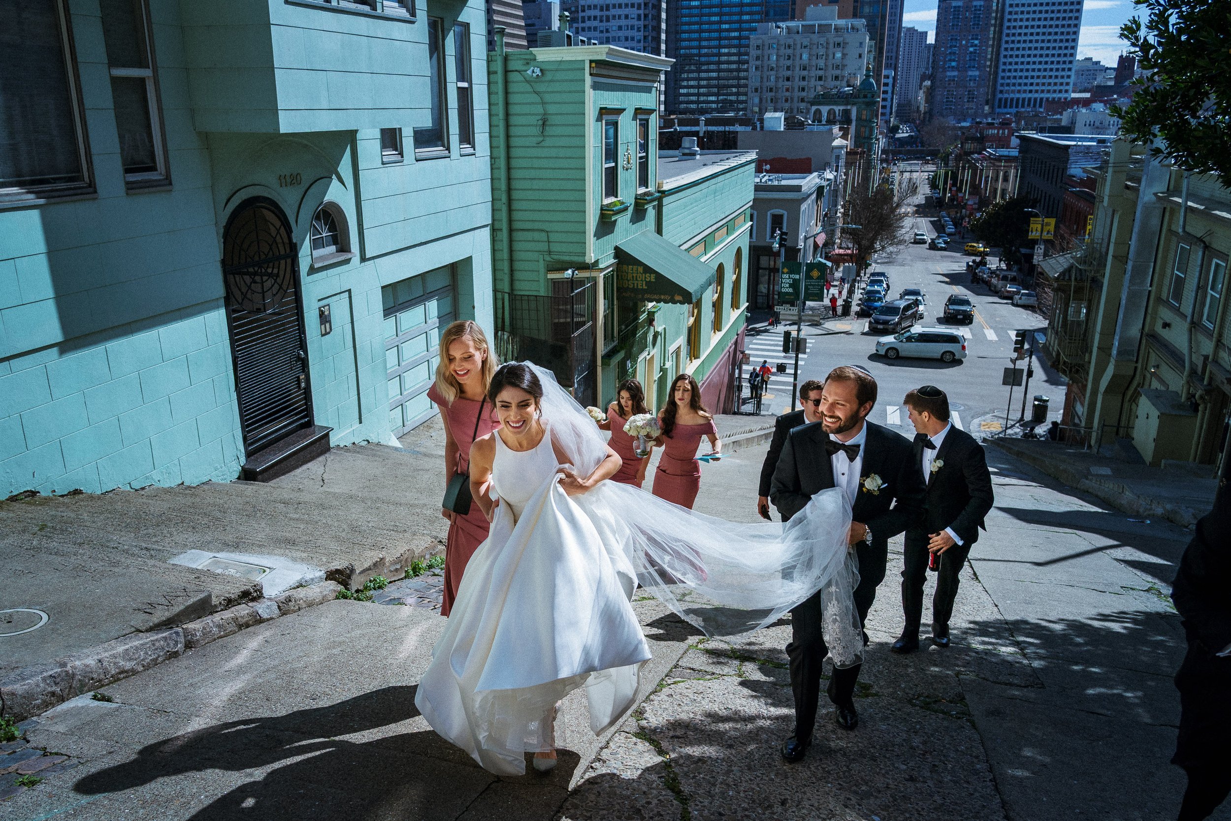 A bride in a white wedding dress and veil, and a groom in a tuxedo, walking up a steep city street with bridesmaids in pink dresses following behind.