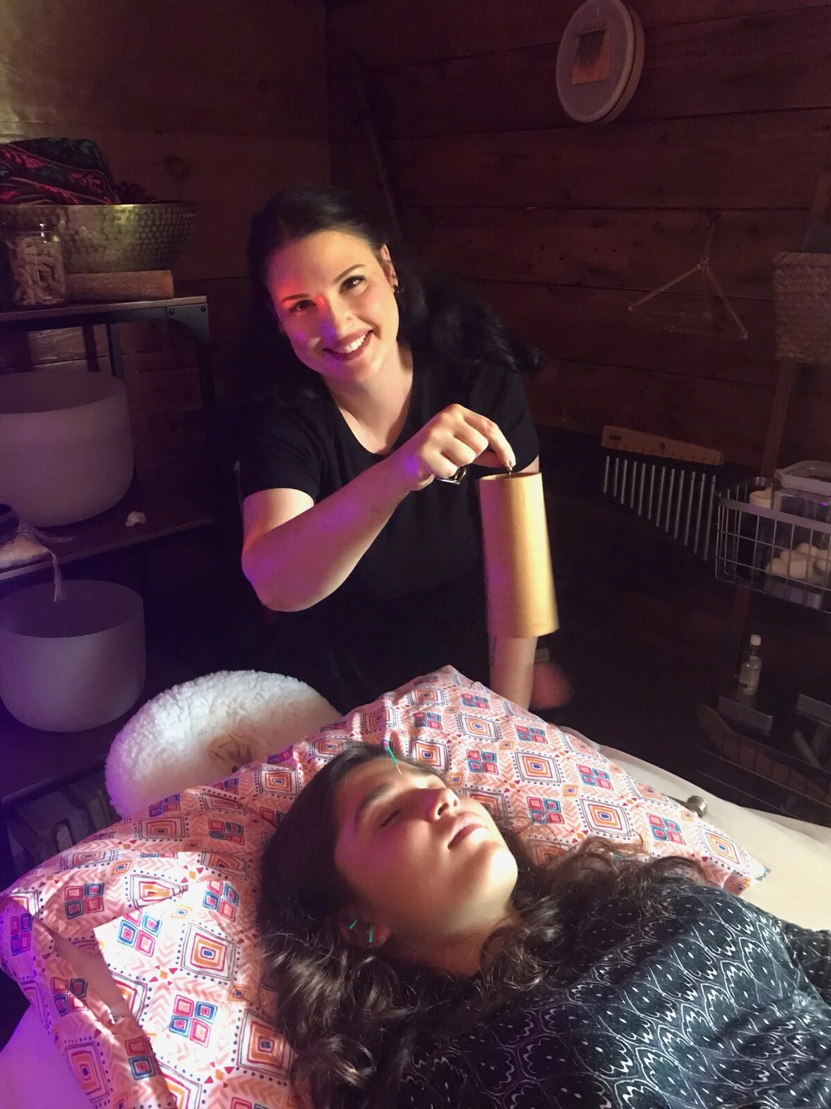 ALT TEXT: Tamara leans over and smiles as she holds a Koshi Chime over a teenage girl who is resting with eyes closed and getting acupuncture.