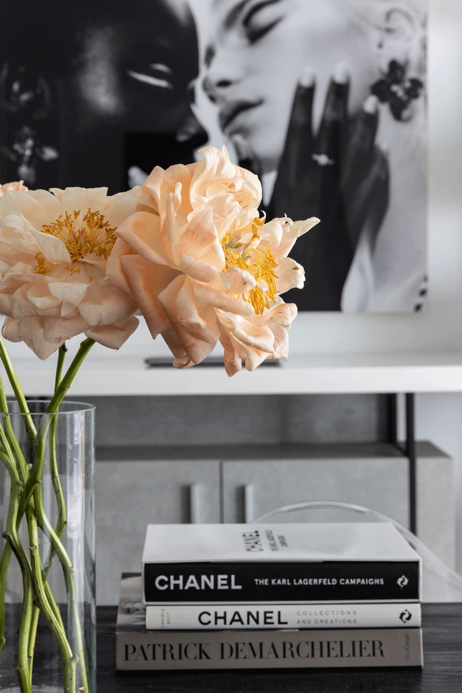 Close-up of peach-colored peony flowers in a glass vase on a table, with a black-and-white portrait of two women in the background and a stack of fashion books including Chanel and Patrick Demarchelier on the table.