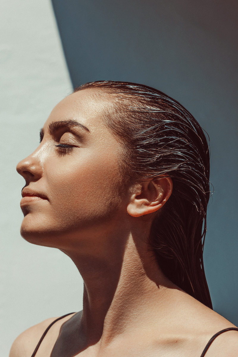 A woman with wet hair, eyes closed, facing sunlight with a peaceful expression.