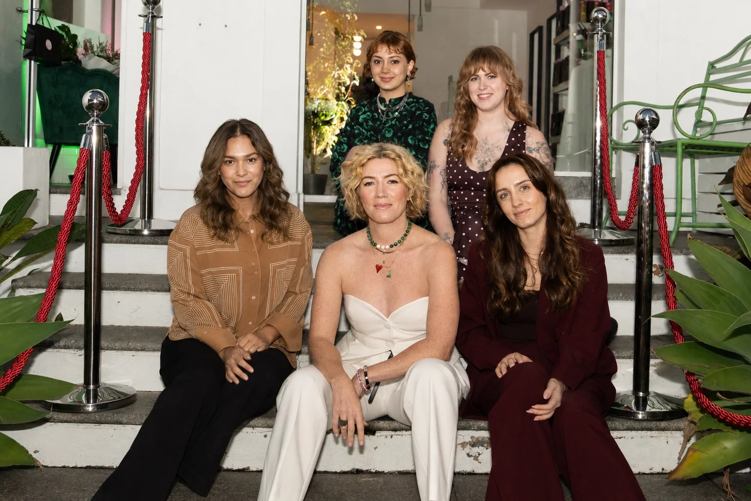 Five women (Loop Hair Salon team) sitting on the steps outside Loop Hair Salon, with plants and a glass door behind them, posing for a group photo.