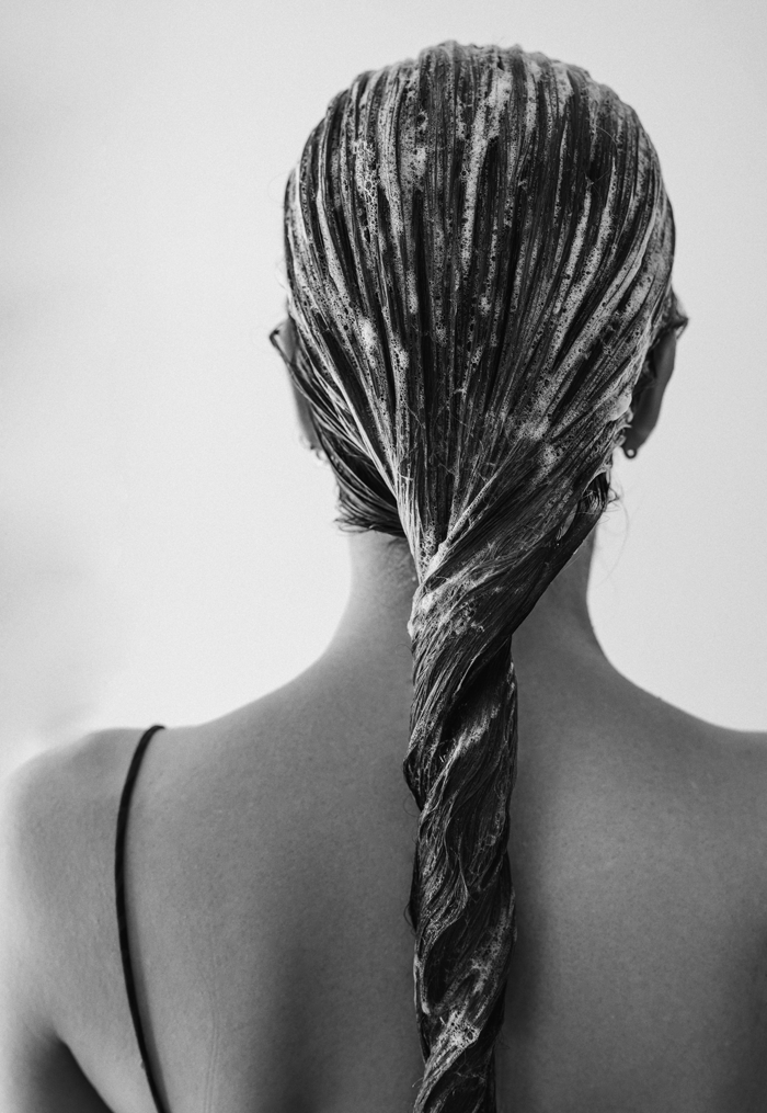 Black and white photo of a woman's back with wet hair twisted into a long braid, with soap or shampoo lather on her hair.