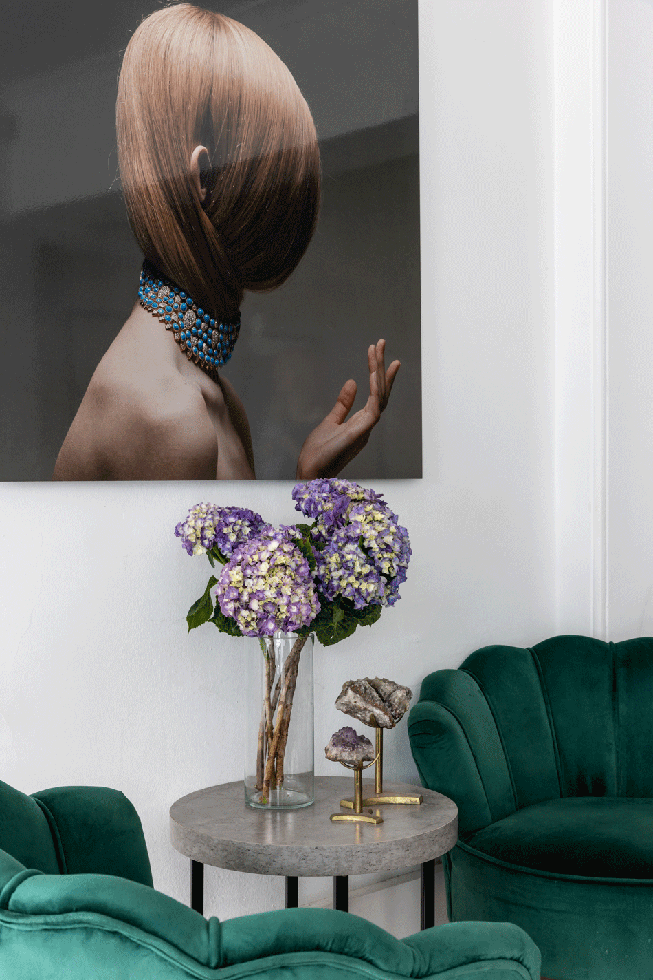 A living room with a large photograph of a woman with a short, auburn hairstyle wearing a blue jeweled choker, looking at a white wall. A round side table with purple hydrangea flowers and mineral or crystal specimens in a glass vase is in front of a green velvet armchair.