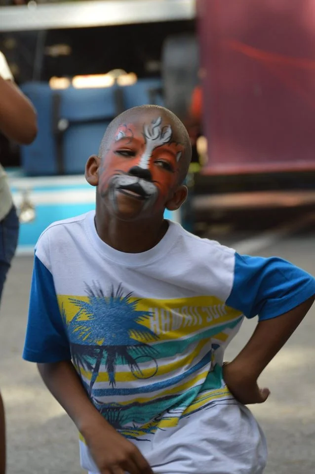 A young boy with face paint resembling a tiger, wearing a white T-shirt with a palm tree and sunset design, standing outdoors with a slight smile and hand on his hip.