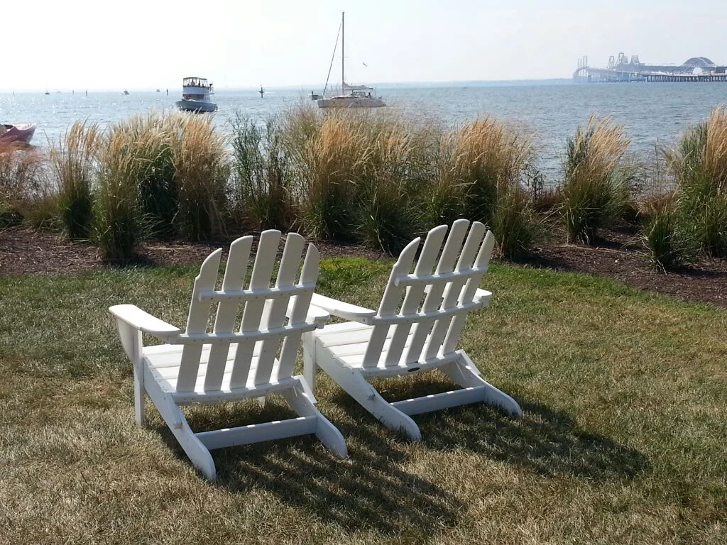 Two white Adirondack chairs facing a body of water with boats and a pier in the distance, surrounded by tall grass and grass on the ground.