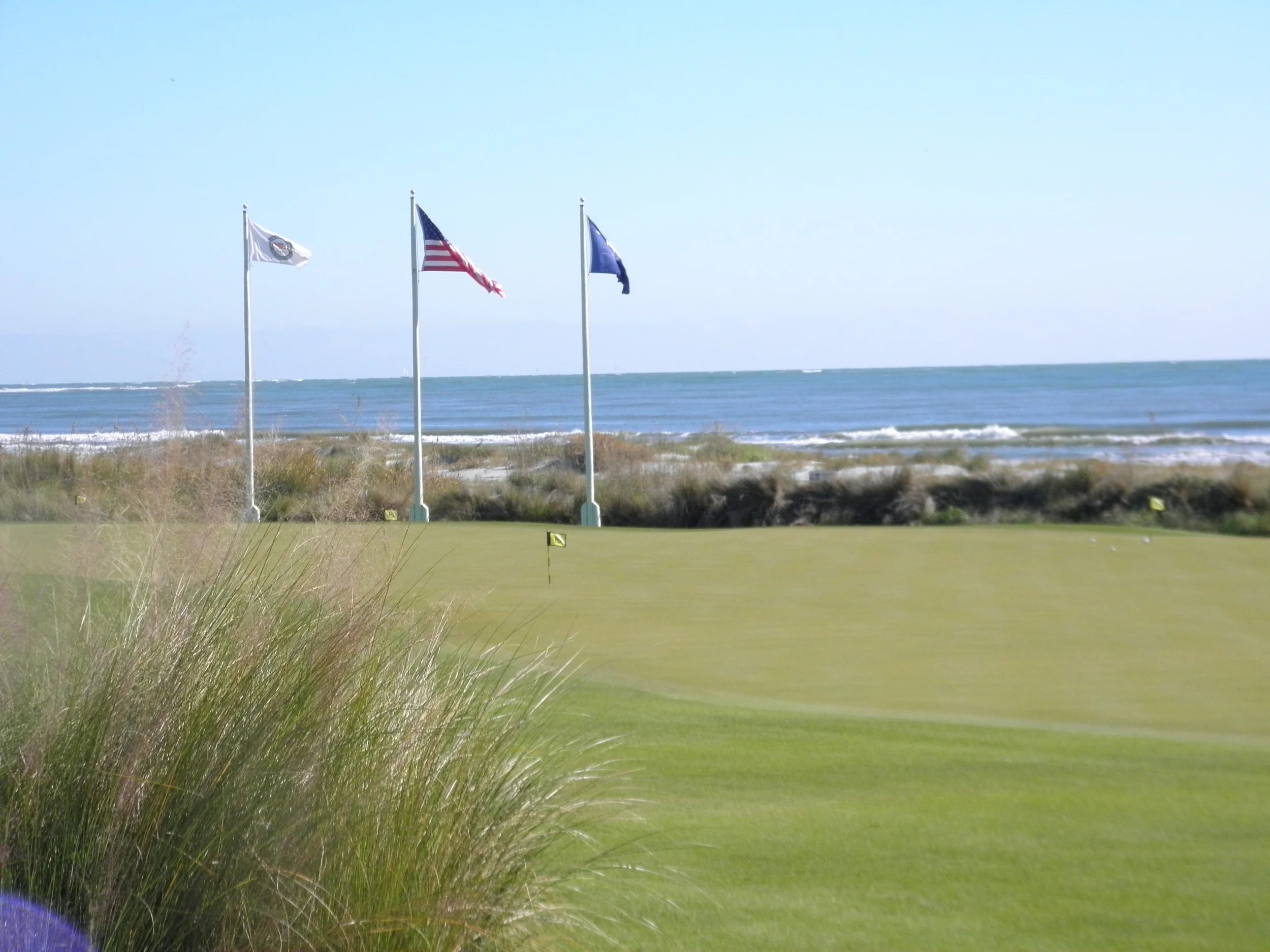 A golf course near the coast with three flags on flagpoles, overlooking the ocean and sandy beach, under a clear blue sky.