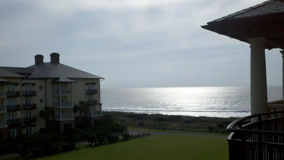 View of the ocean reflecting sunlight, with a grassy area, residential building, and balcony in the foreground.