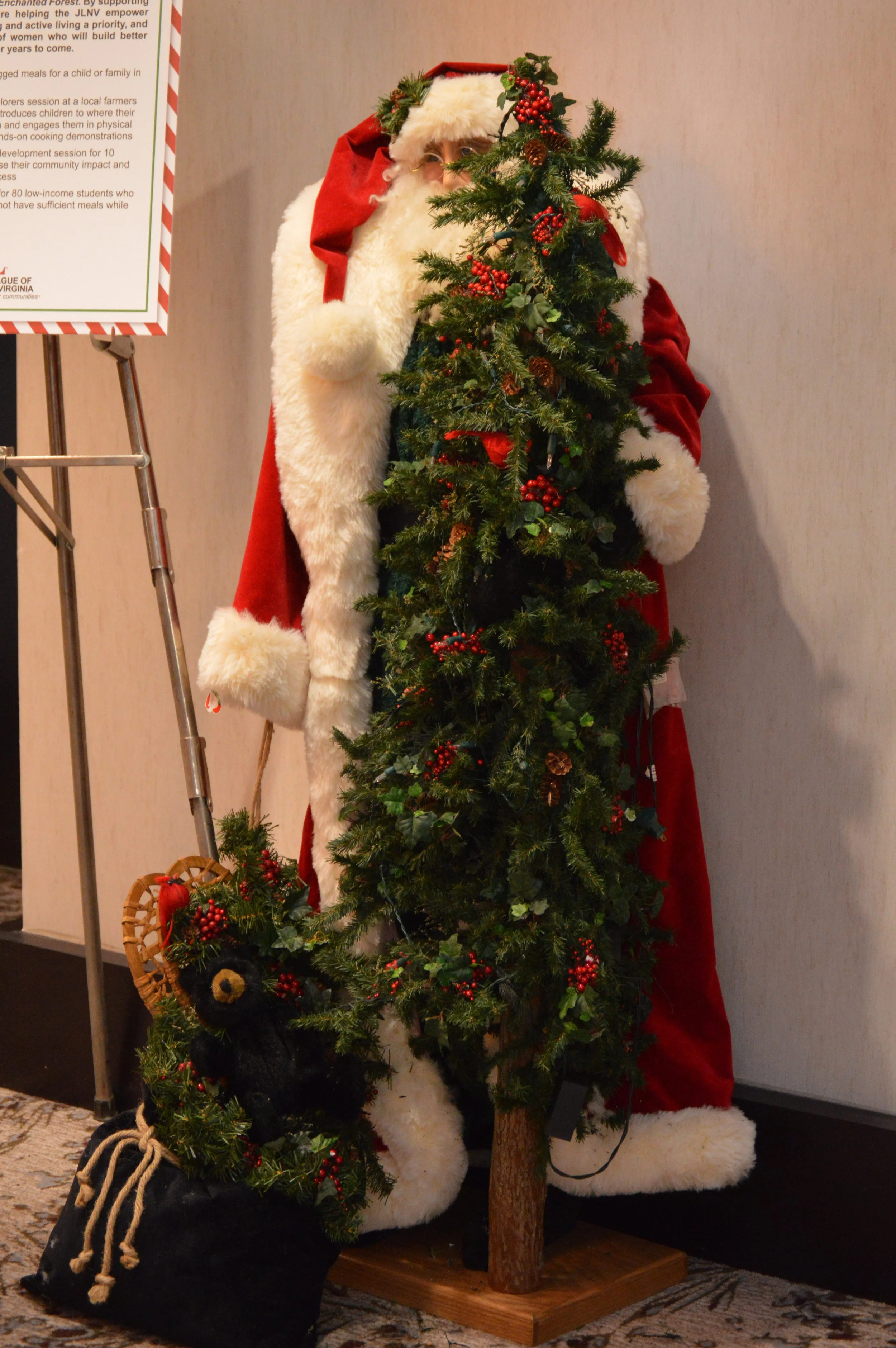 A Santa Claus figure standing next to a decorated Christmas tree with red berries and pinecones. Santa is dressed in a red coat with white fur trim, and a matching red hat with white fur trim. A small black teddy bear ornament is in a black bag fille