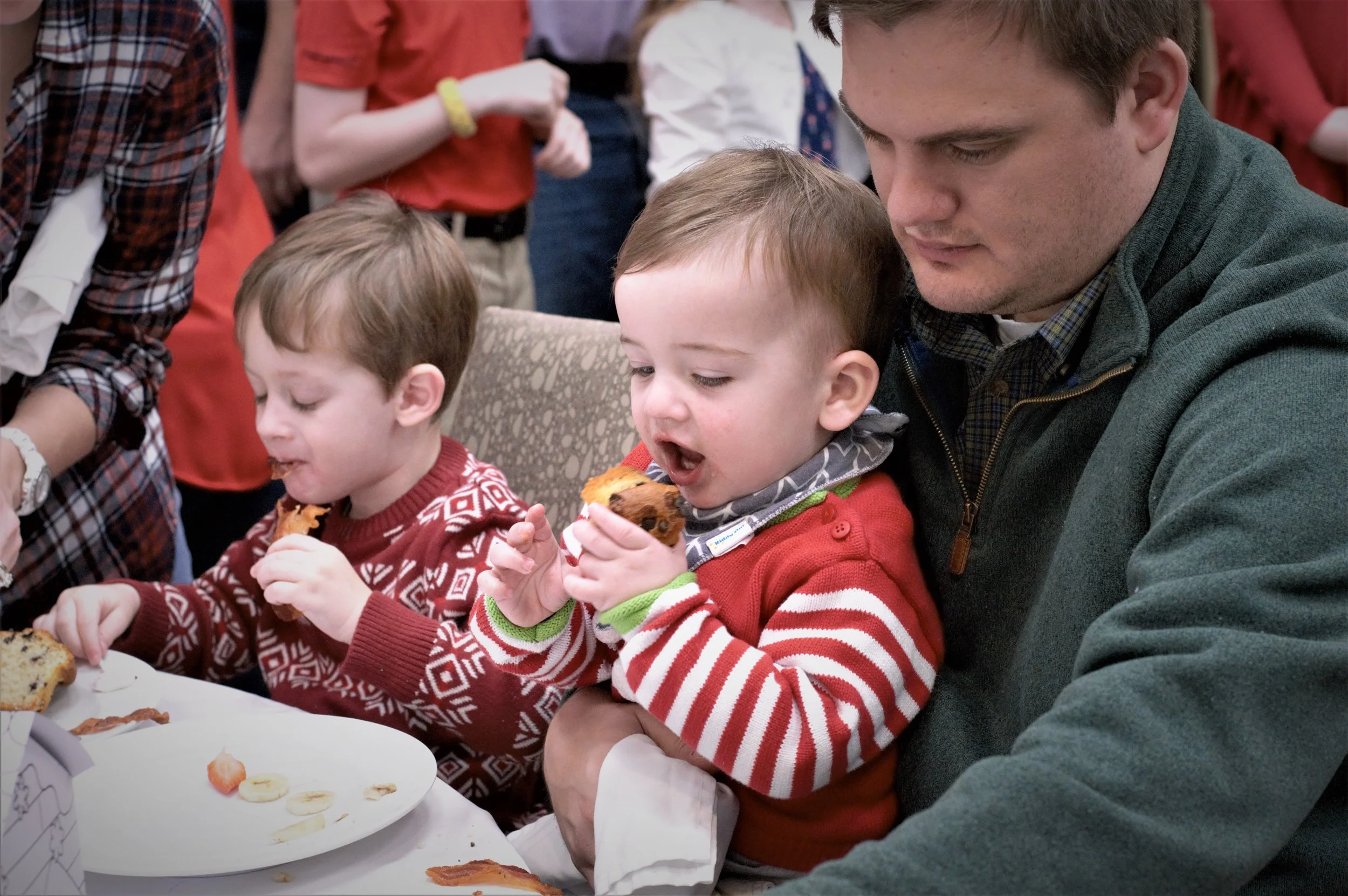 A man and two young boys sitting at a table eating slices of cake and bananas, with people standing in the background.