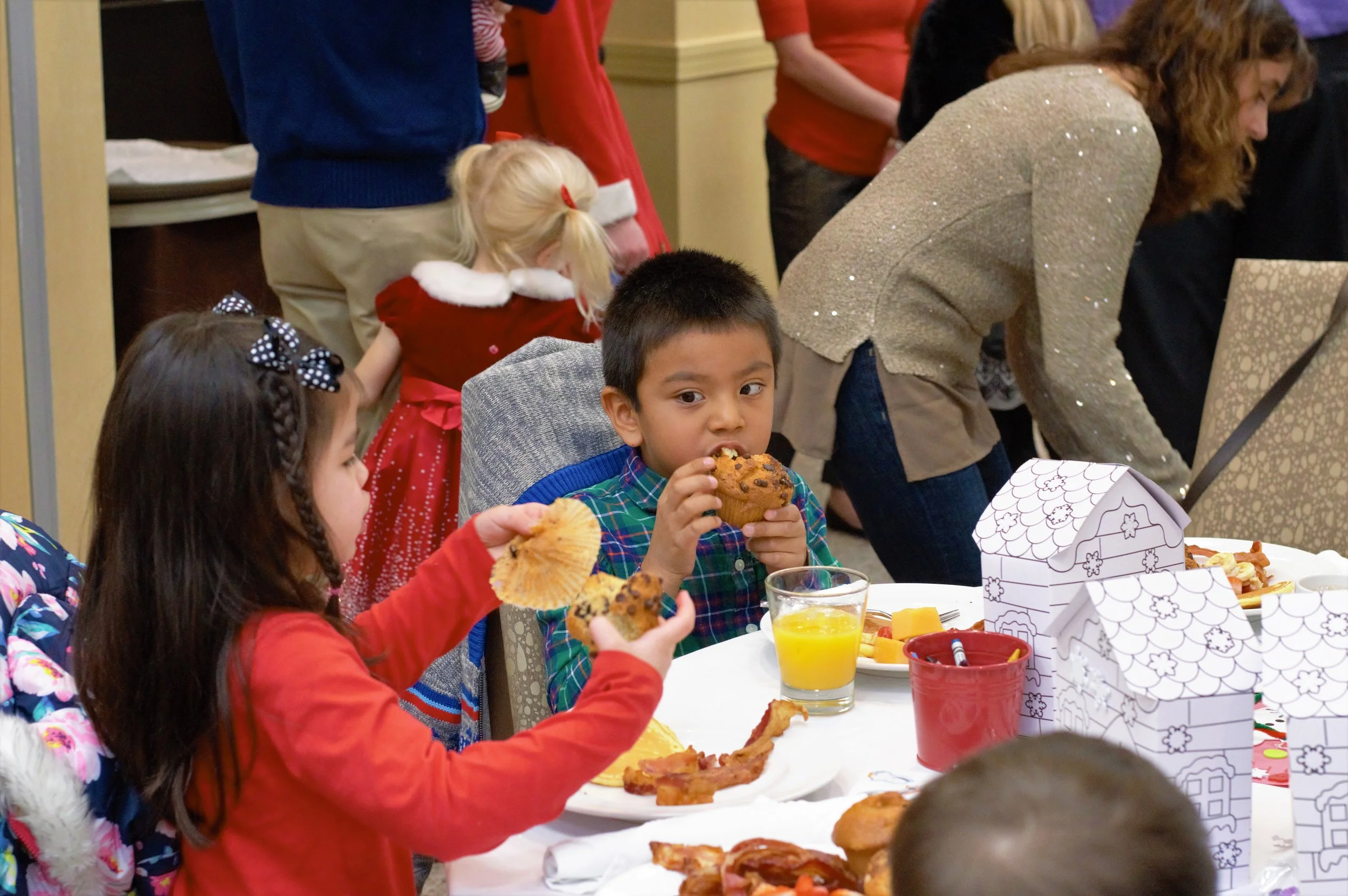 Children sitting at a table eating breakfast, with festive food and drinks, and adults and children in the background.