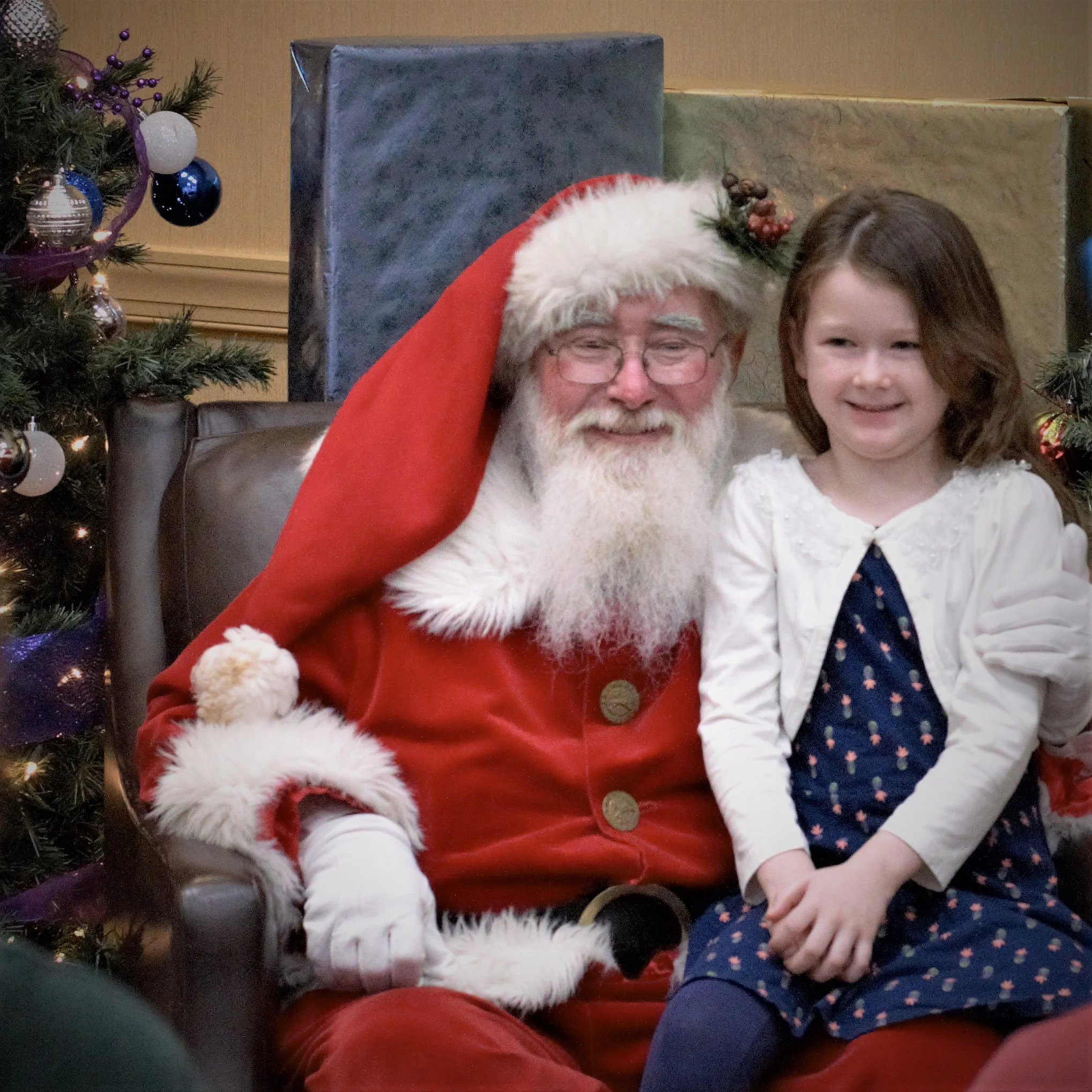 Santa Claus with white beard and glasses sitting next to a young girl in front of a Christmas tree and large wrapped presents.