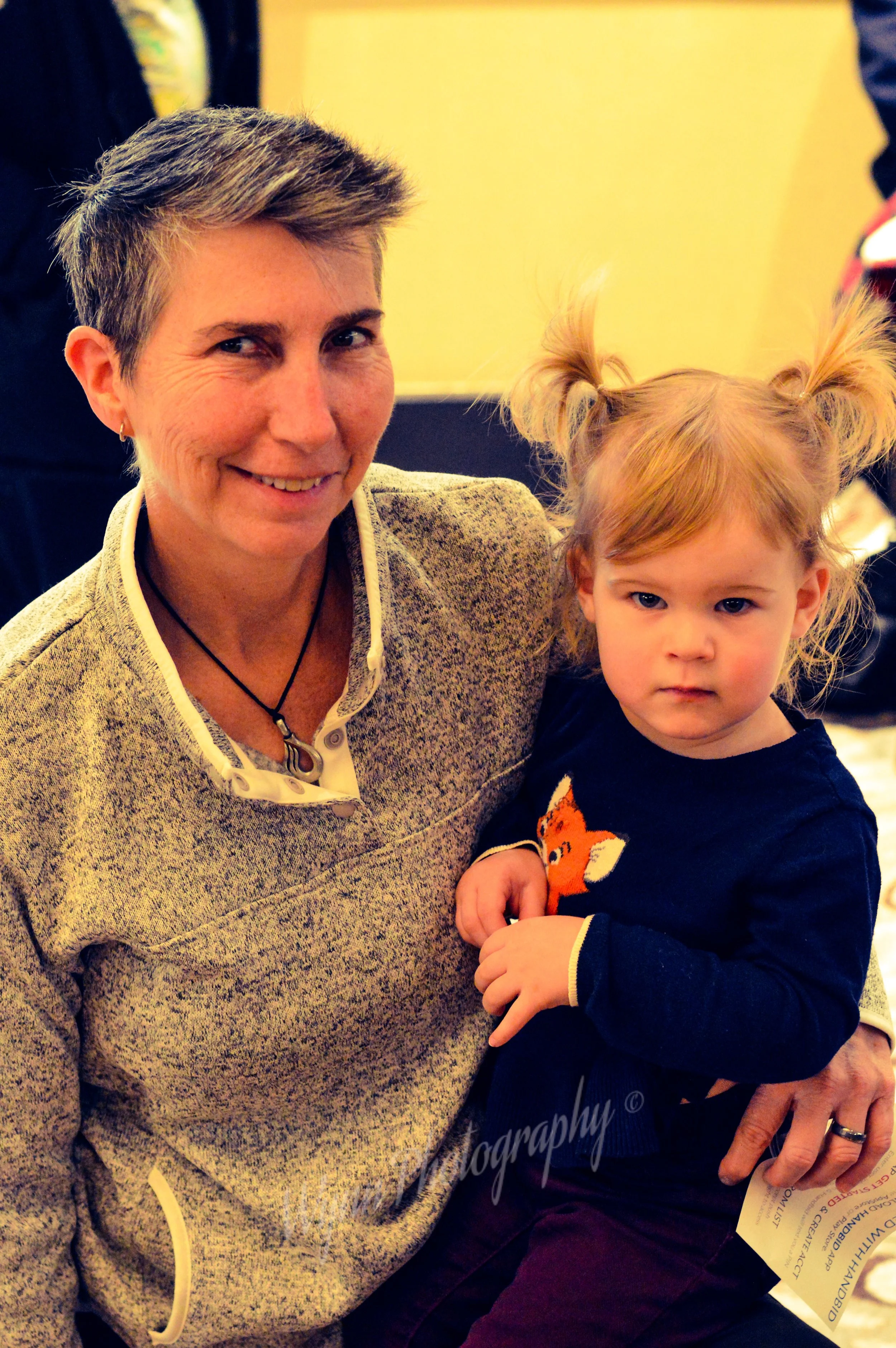 A woman with short gray hair smiling while holding a young girl with blonde hair tied in pigtails. The woman wears a beige sweater and the girl wears a black shirt with a fox design.