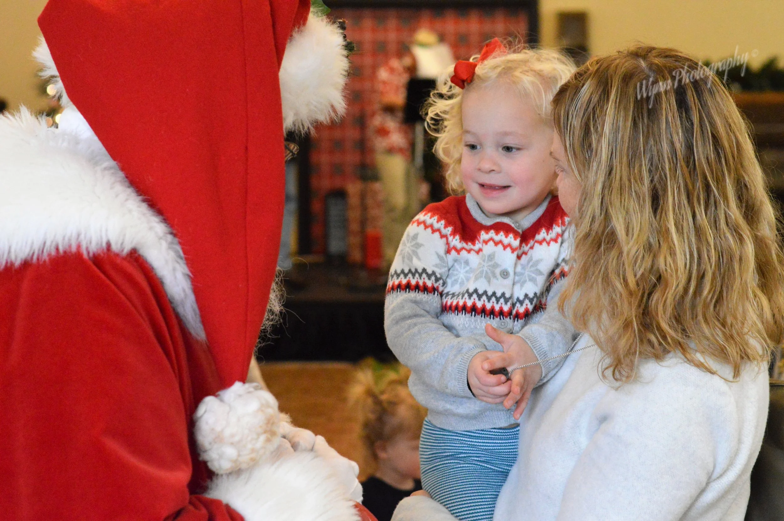 A young girl with curly blonde hair and a woman, possibly her mother, are smiling while talking to a person dressed as Santa Claus. The girl is wearing a festive sweater, and the woman has long, wavy hair. Santa's red suit and white beard are partial