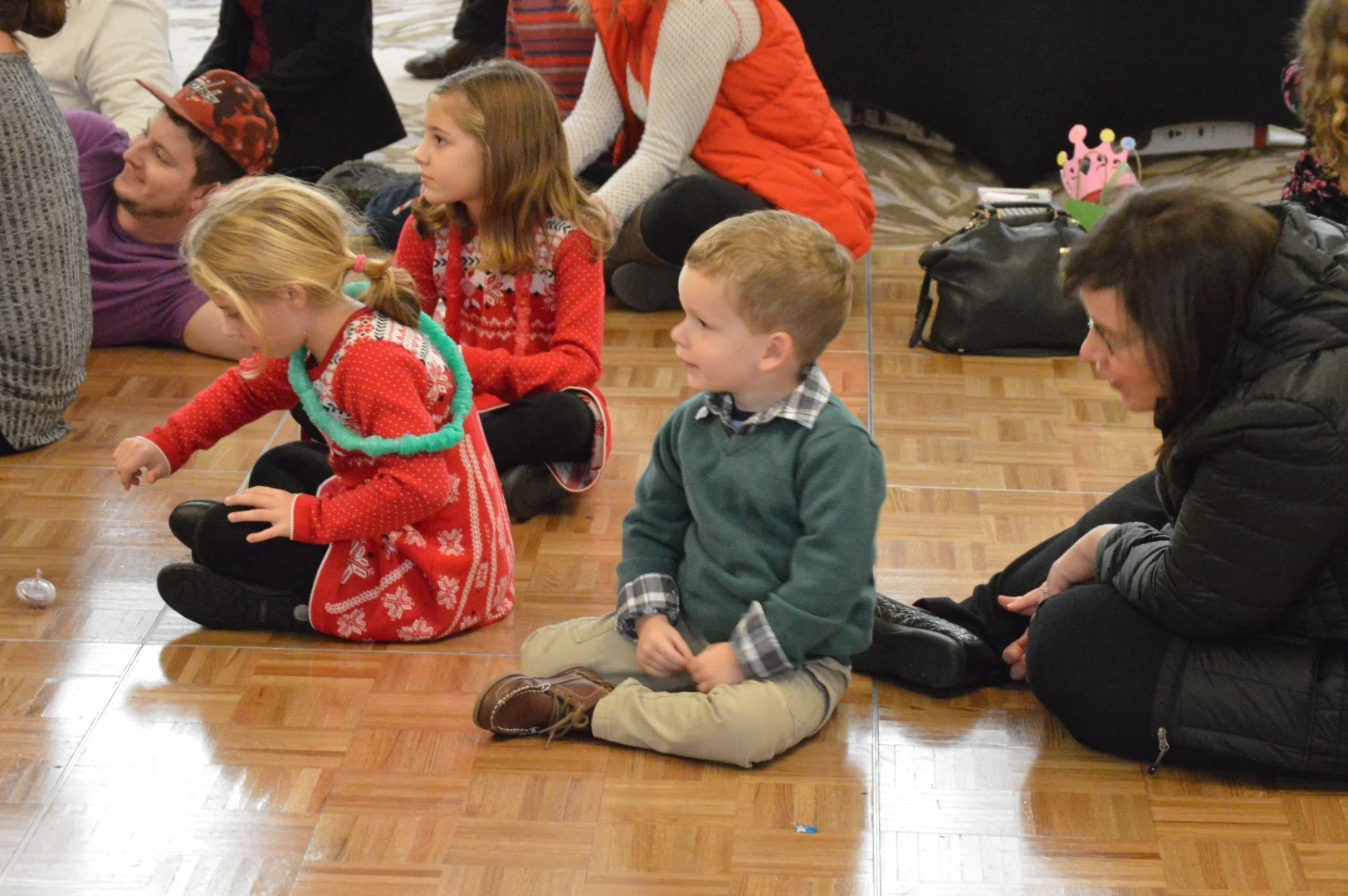 Children and adults sitting on a wooden floor, participating in an indoor event, with some children dressed in holiday clothing.