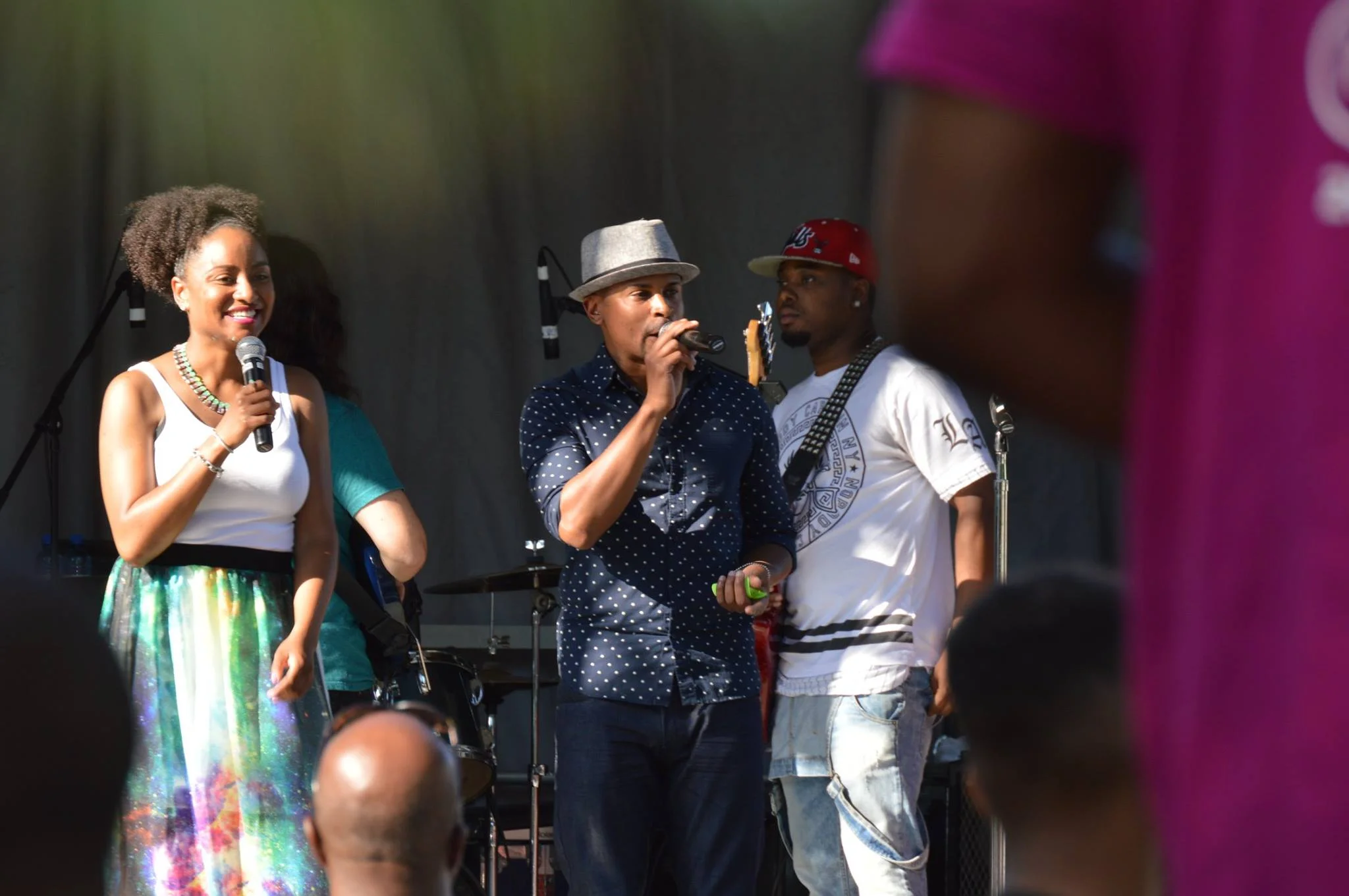 Group of people on stage during outdoor event, woman smiling into microphone, man in polka dot shirt with microphone, man in white T-shirt and red cap, musical instruments in background.
