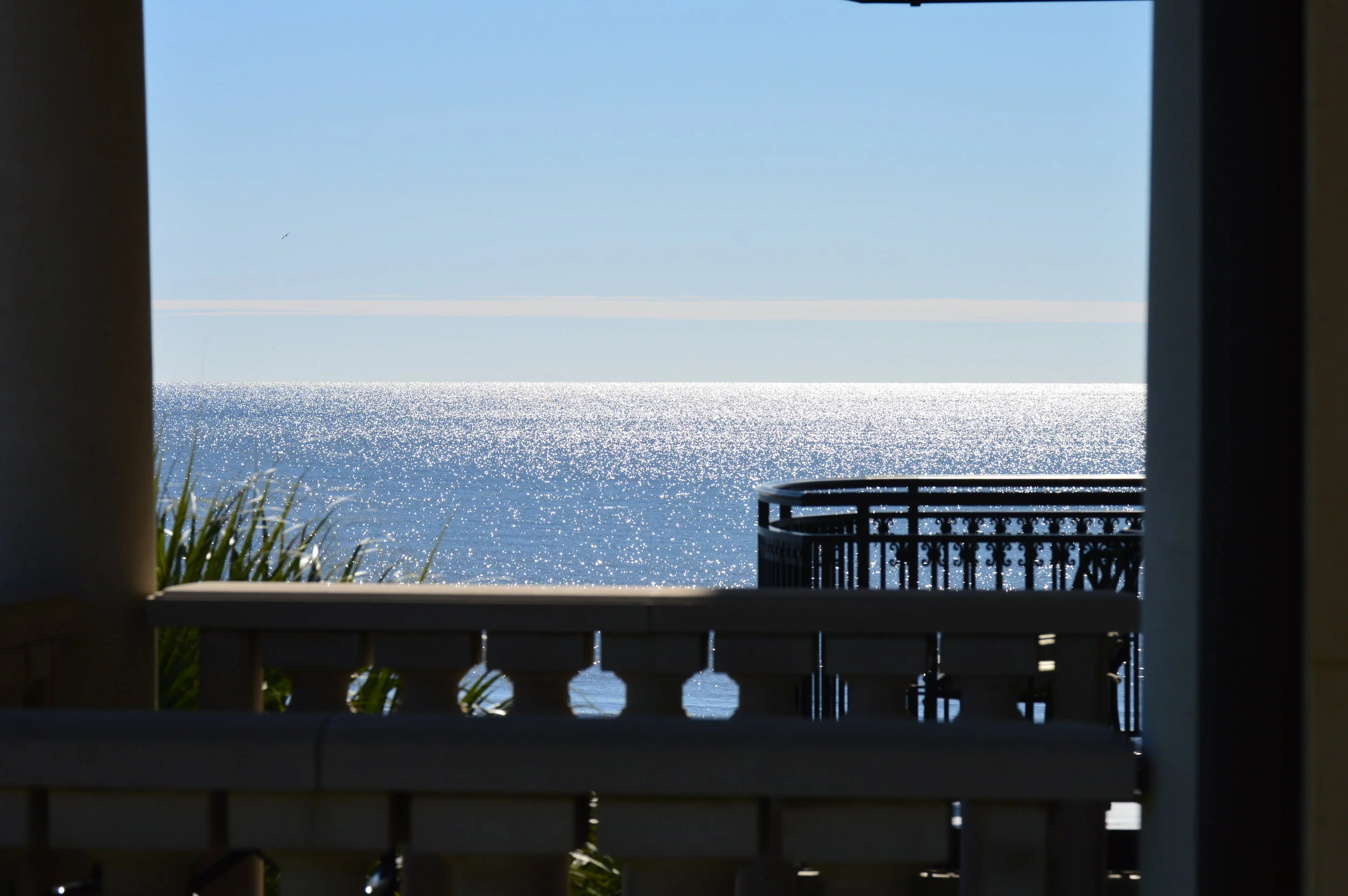 View of the ocean through a window with a balcony railing seen in the foreground, sparkling water under a clear blue sky.