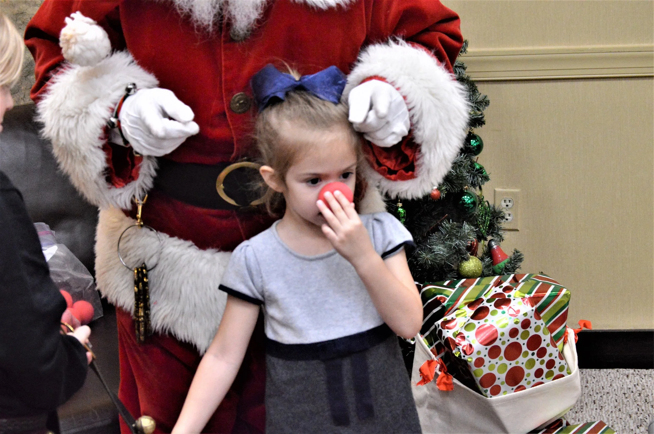 A young girl with a blue bow in her hair and a gray dress with black trim stands in front of Santa Claus, who is wearing a red velvet coat with white fur trim. Santa is holding the girl's head and standing next to a Christmas tree decorated with gree
