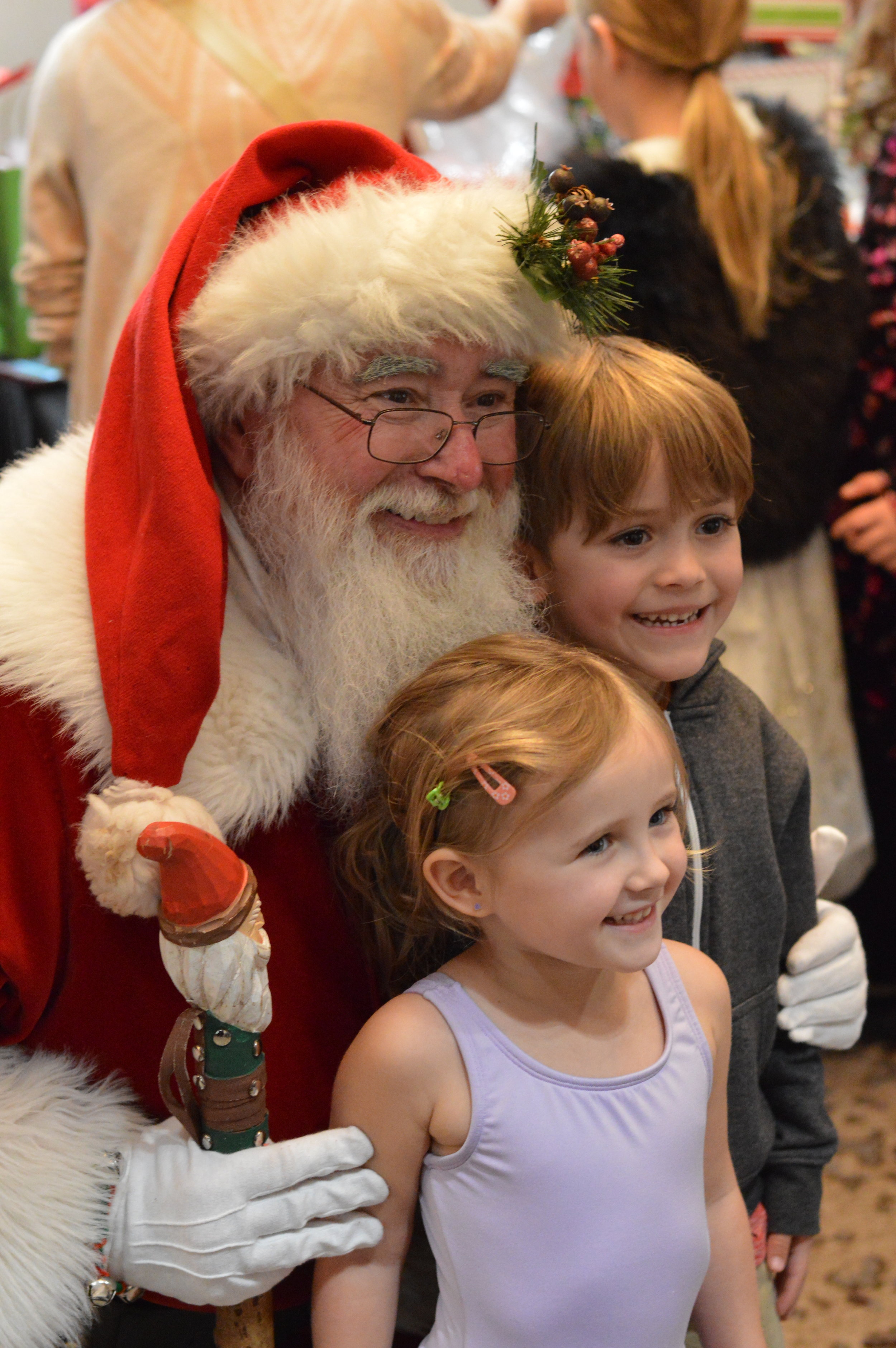Santa Claus in a red suit with white fur trim, wearing glasses, holding a staff, taking a photo with smiling children at a holiday event.