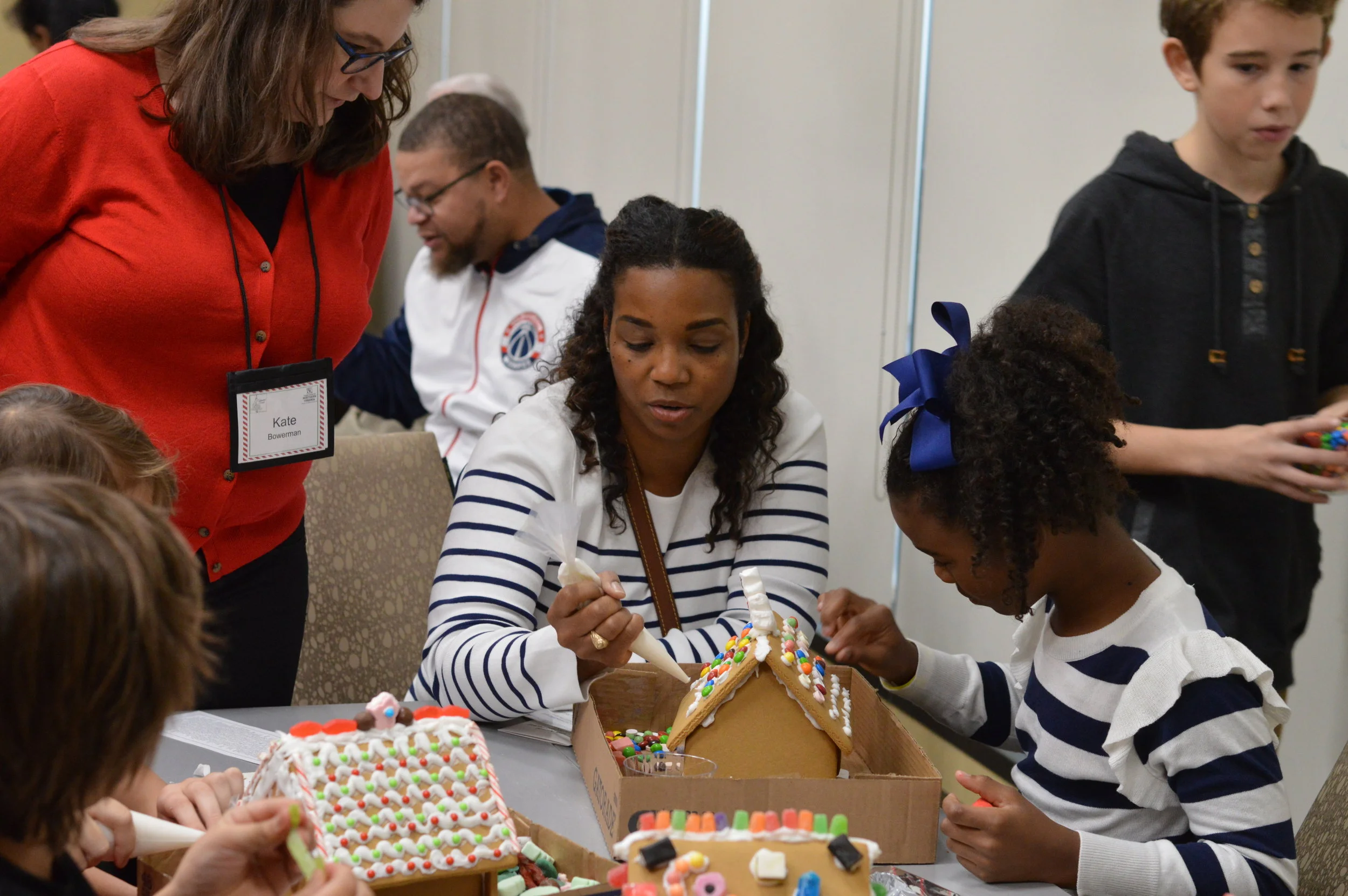 A woman in a striped shirt and a young girl with a blue bow decorating gingerbread houses at a table, while other children and adults stand nearby. The woman is using a piping bag to add icing to the gingerbread house.
