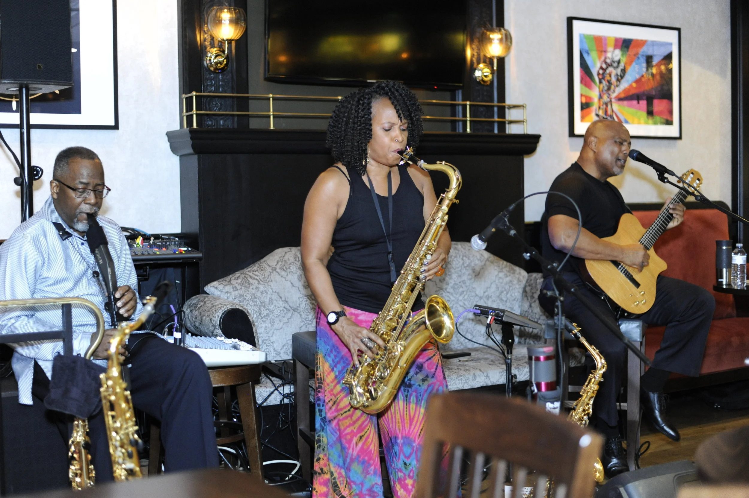Group of three jazz musicians performing indoors, with a woman playing saxophone in the center, a man playing keyboard on the left, and another man playing acoustic guitar on the right, with colorful artwork on the wall behind them.