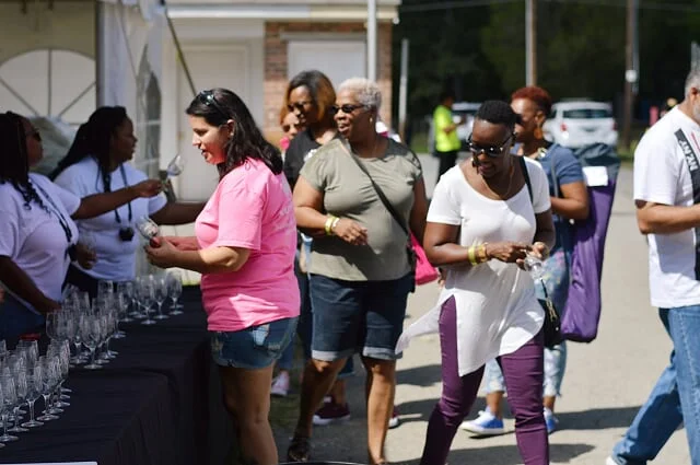 People at an outdoor event, some choosing glasses from a table with wine glasses, under a tent.