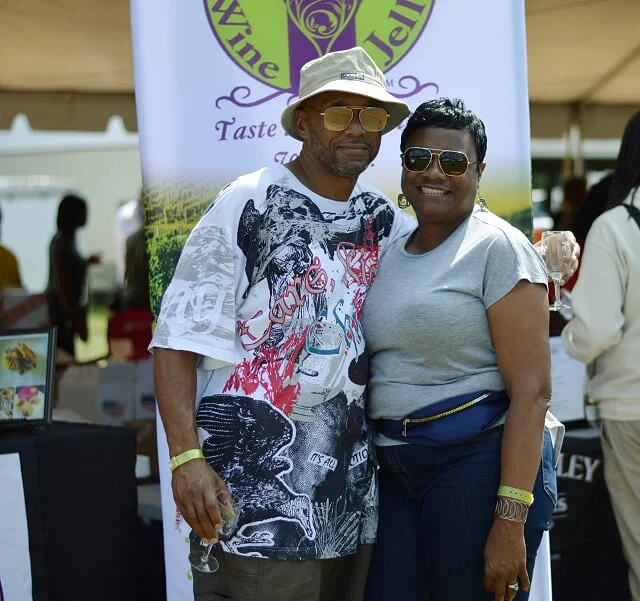 Two people smiling and posing at an outdoor event, standing in front of a banner related to wine and jelly.