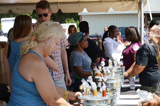 People gathered outdoors under a tent at a table with various bottles and containers, likely at a vendor booth or festival.