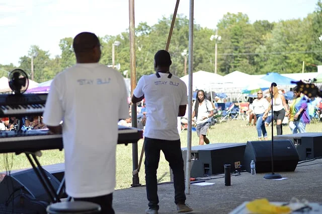 Musicians performing on an outdoor stage with a crowd of people dancing and enjoying the event under tents and purple umbrellas.