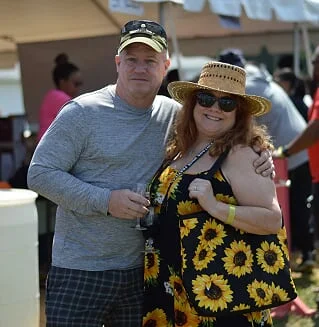 A man and a woman stand together outdoors, smiling. The woman wears a sunflower-patterned dress and a wide-brimmed straw hat, holding a camera, with other people and tents visible in the background.