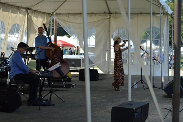 Musicians performing on a stage under a white tent at an outdoor event, including a pianist, a double bassist, and a female vocalist wearing a hat.