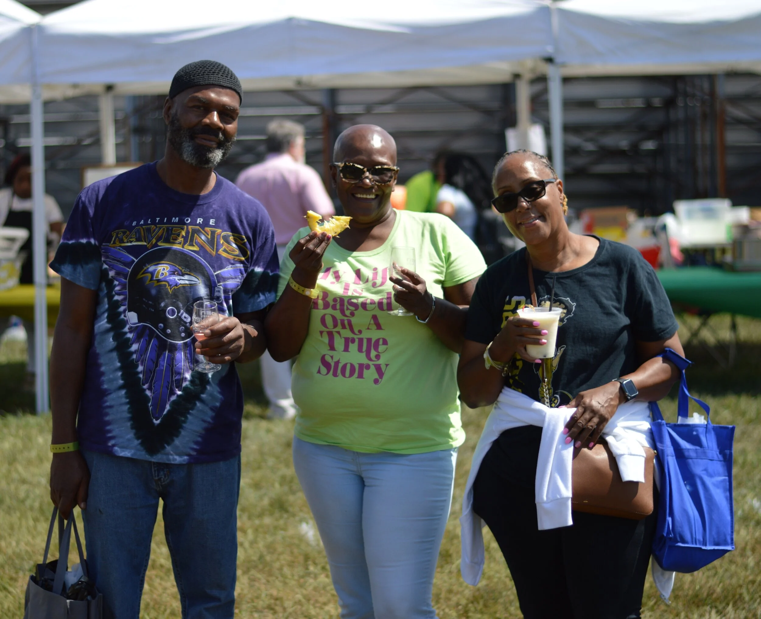 Three middle-aged people standing together outdoors at a sunny event, smiling and holding snacks and drinks, with tents and other people in the background.