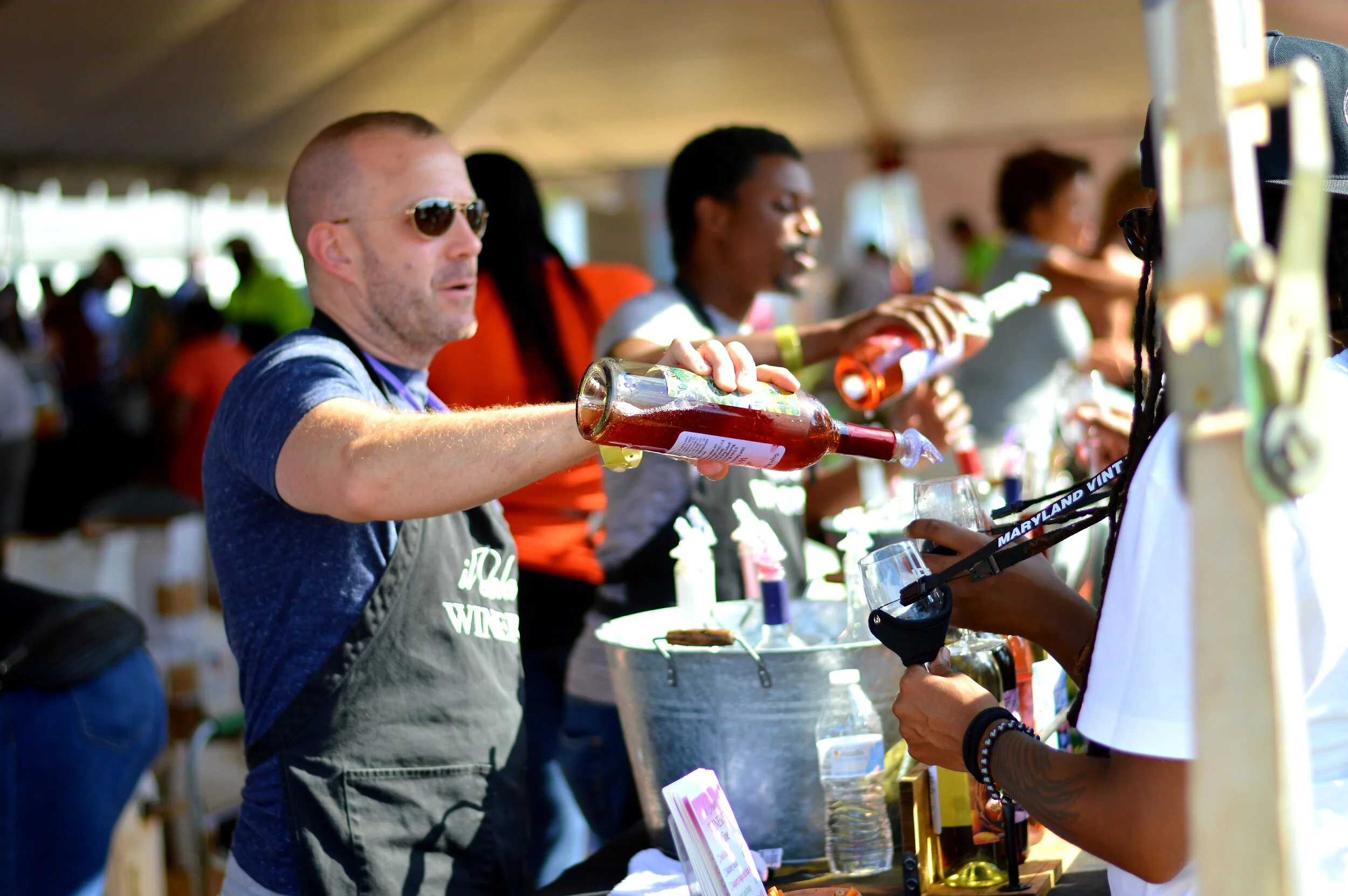 A man wearing sunglasses and a gray apron is pouring a red drink into a glass at an outdoor event, with several other people in the background also preparing drinks.