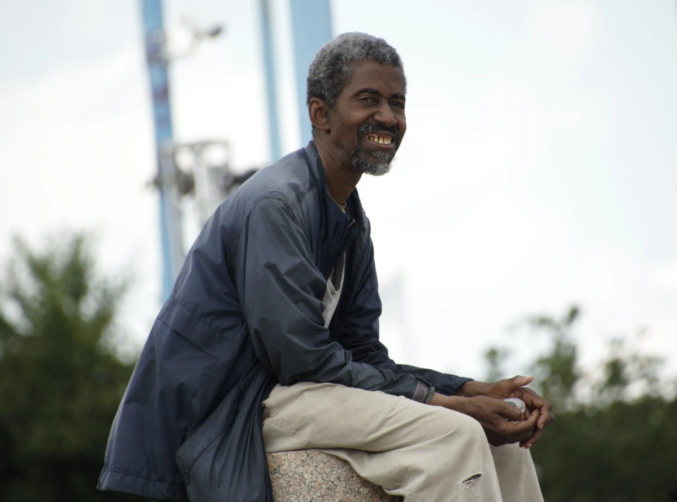 A smiling man with gray hair and a beard, sitting outdoors on a stone surface, wearing a dark jacket and light-colored pants.