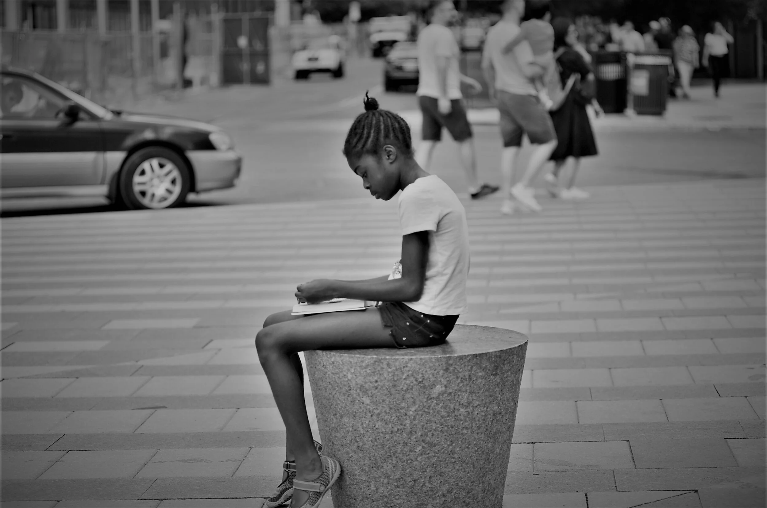 A young girl with braided hair sits on a cylindrical stone bench in an urban setting, reading or writing in a notebook. In the background, several pedestrians walk by on the sidewalk, and a car is parked on the street.
